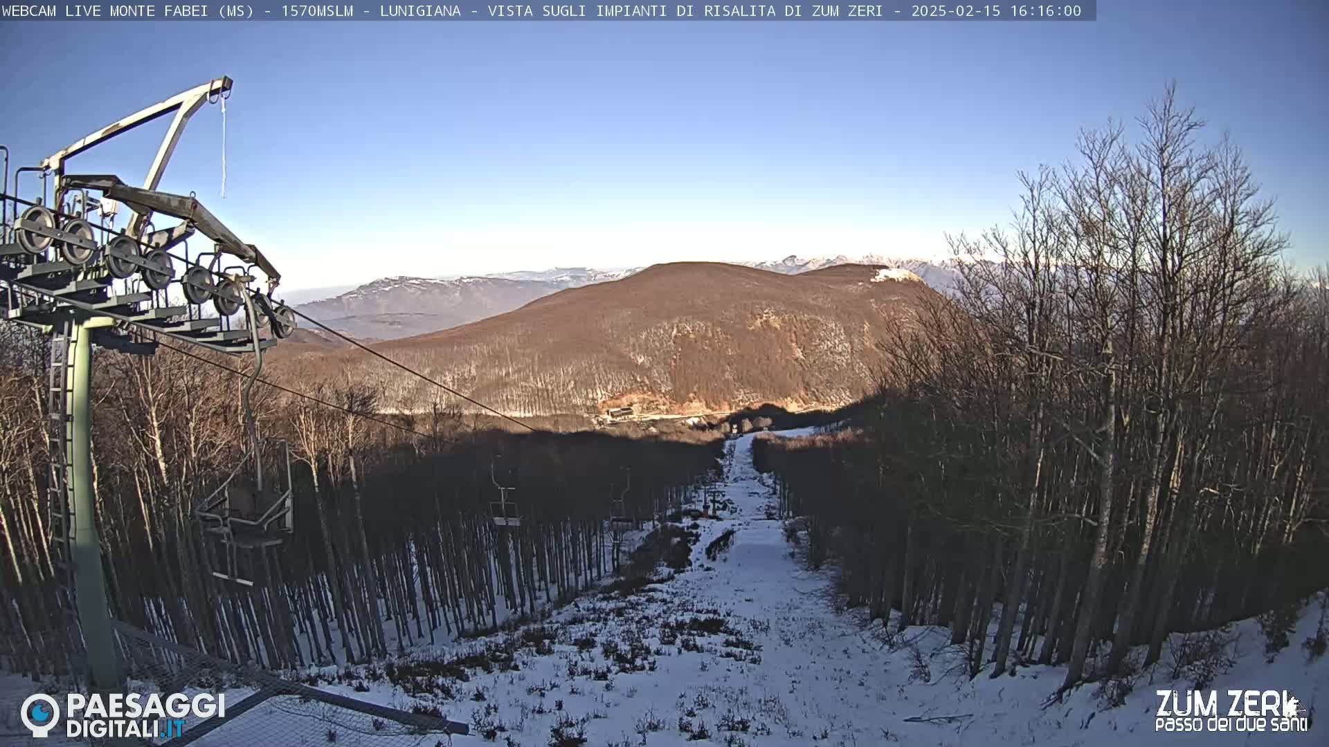 A ski lift ascends a snow-covered hill through a bare, winter forest under a clear blue sky.
