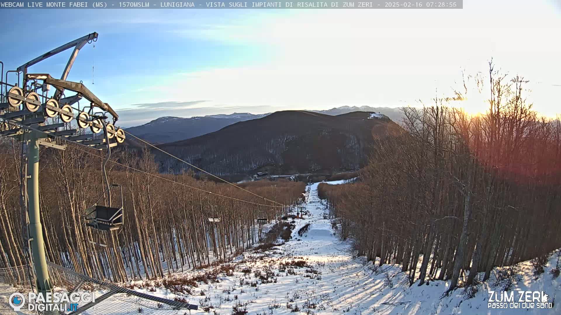 A ski lift ascends a snow-covered mountain slope under a partly sunny sky at sunrise, surrounded by leafless trees.