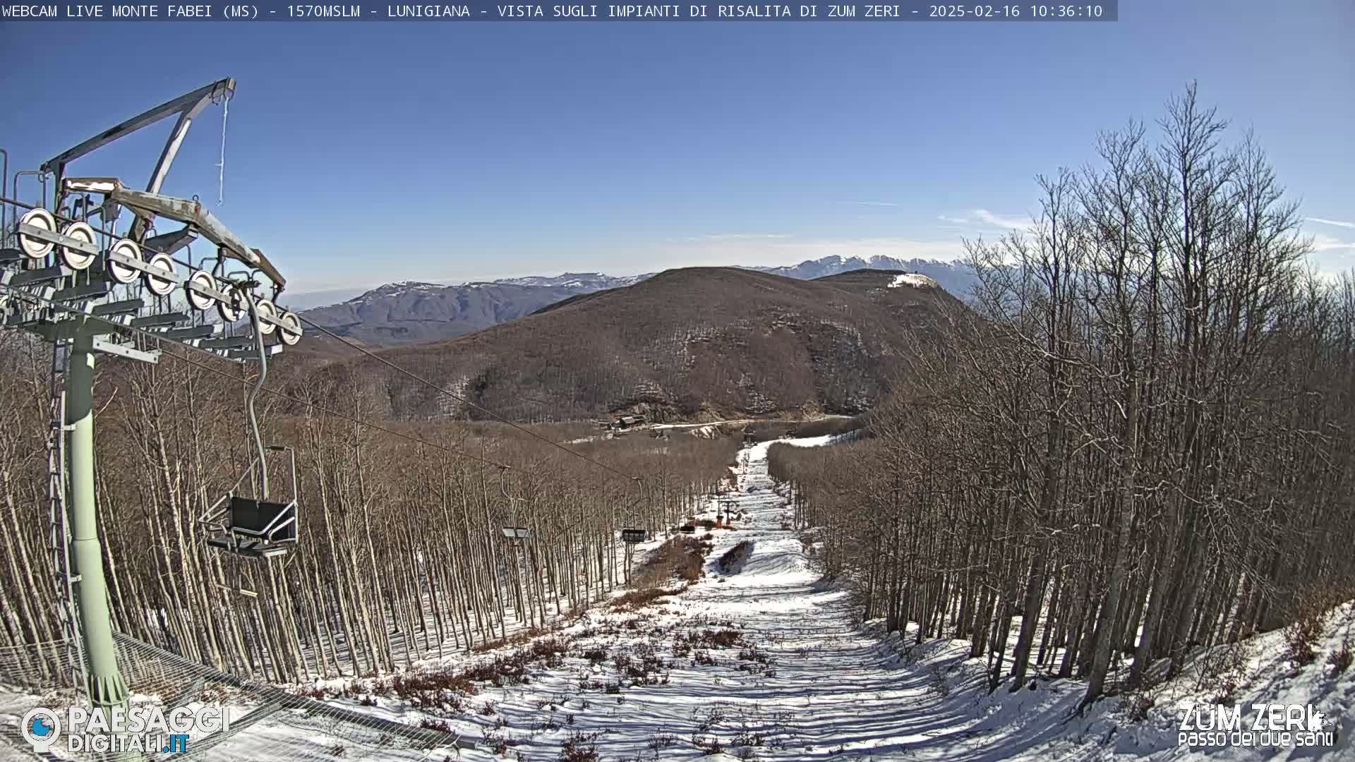 A ski lift chair and cable ascend a snowy slope through a sparsely snow-covered forest on a sunny day, with mountains in the background.