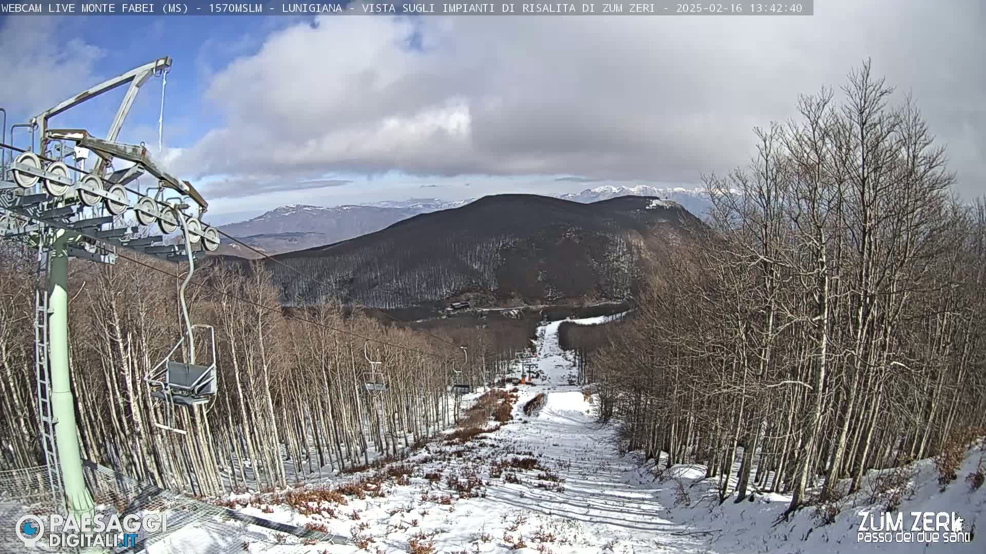 A snowy ski slope descends through a forest of bare trees under a partly cloudy sky, with mountains in the background.