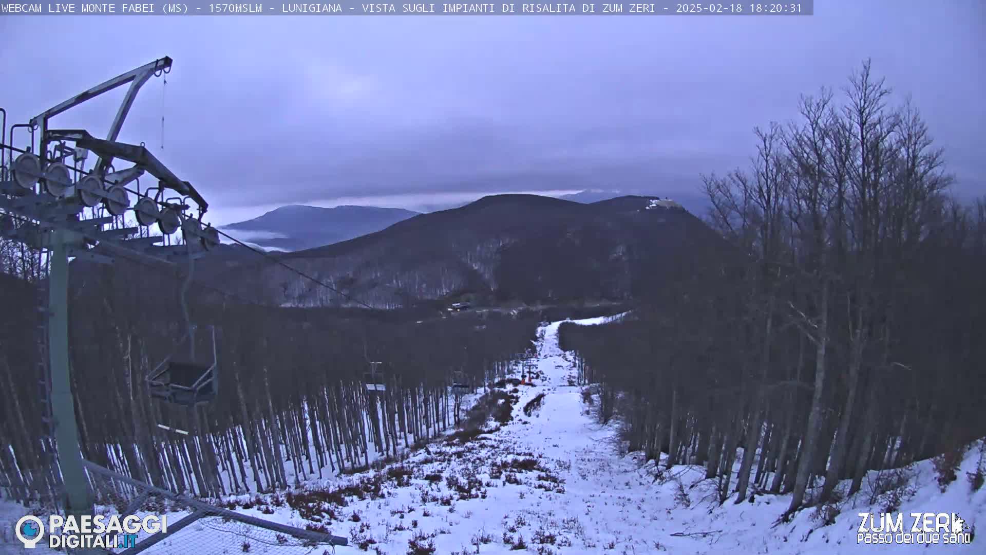 A snow-covered ski slope descends from a mountaintop, with a ski lift visible in the foreground under an overcast sky.