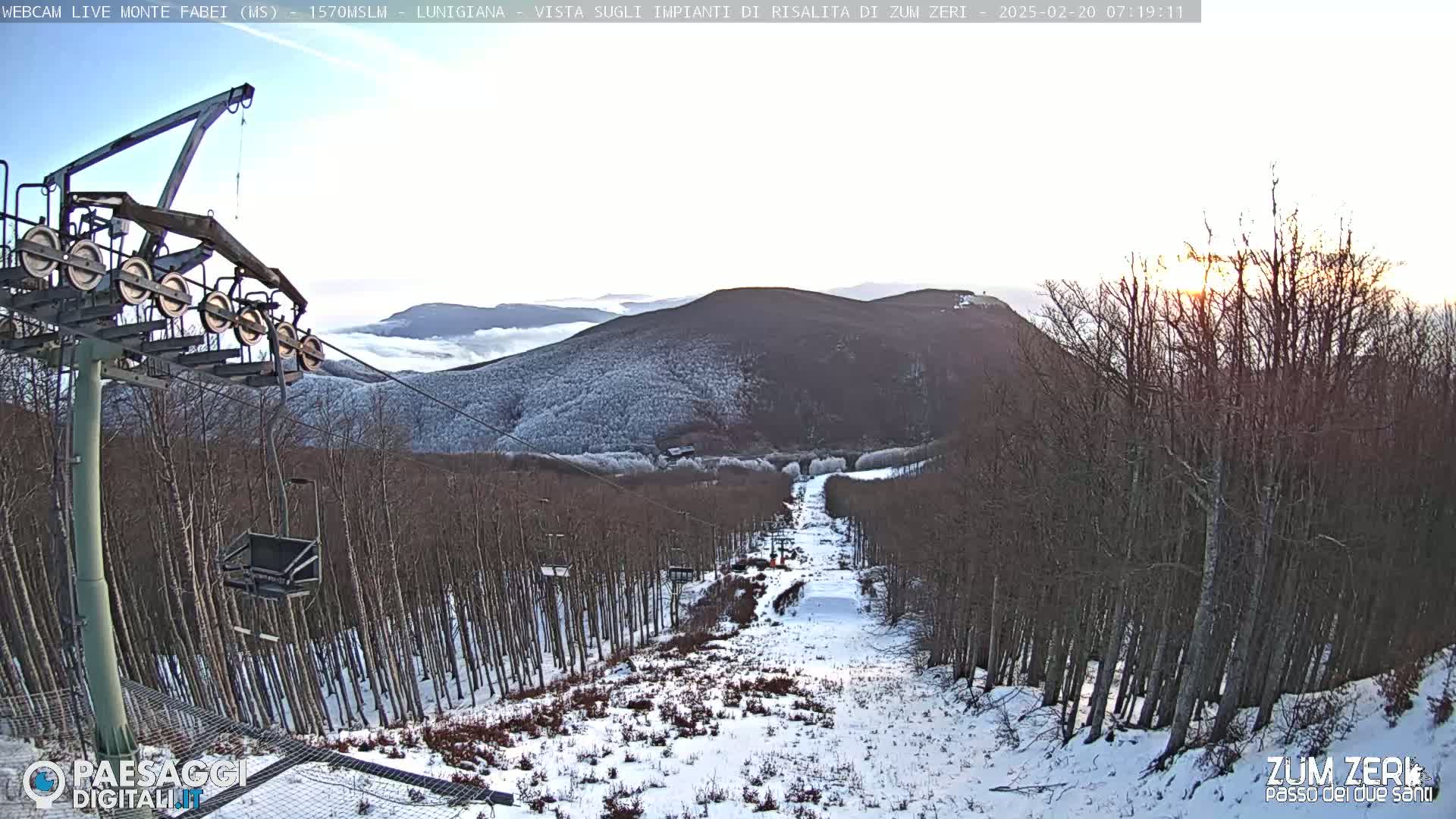 A snowy ski slope with a chairlift and bare trees is viewed at sunrise, with a layer of fog in the distant valley.