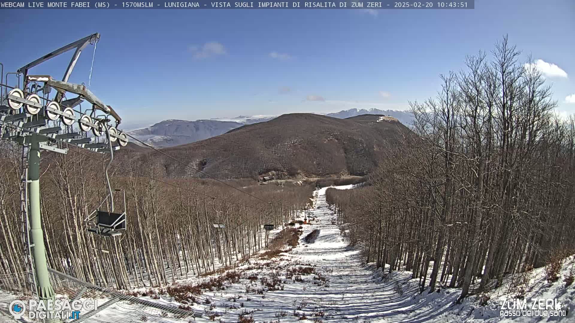 A ski lift chair ascends a snowy mountain slope lined with leafless trees under a mostly clear blue sky.