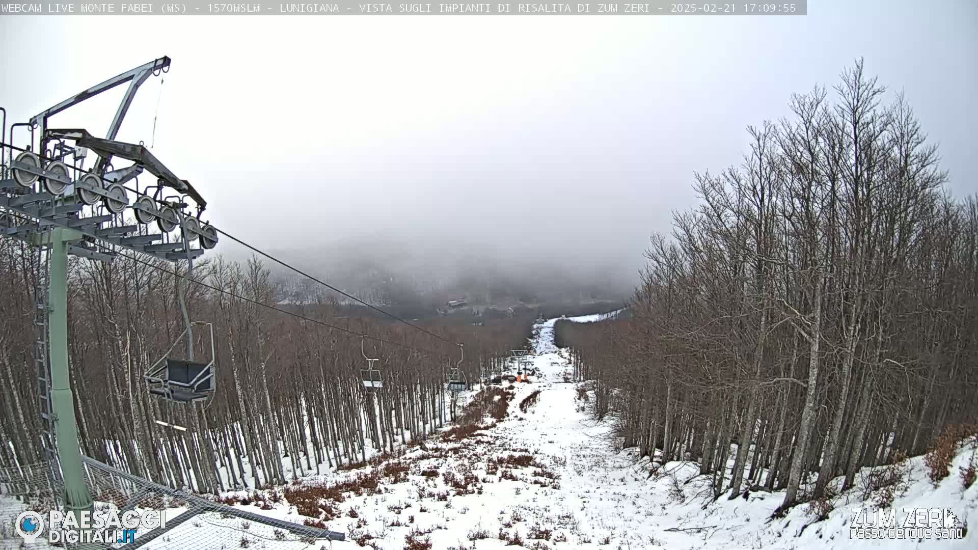 A snow-covered ski slope descends through a foggy, leafless forest, with a chairlift visible in the foreground.