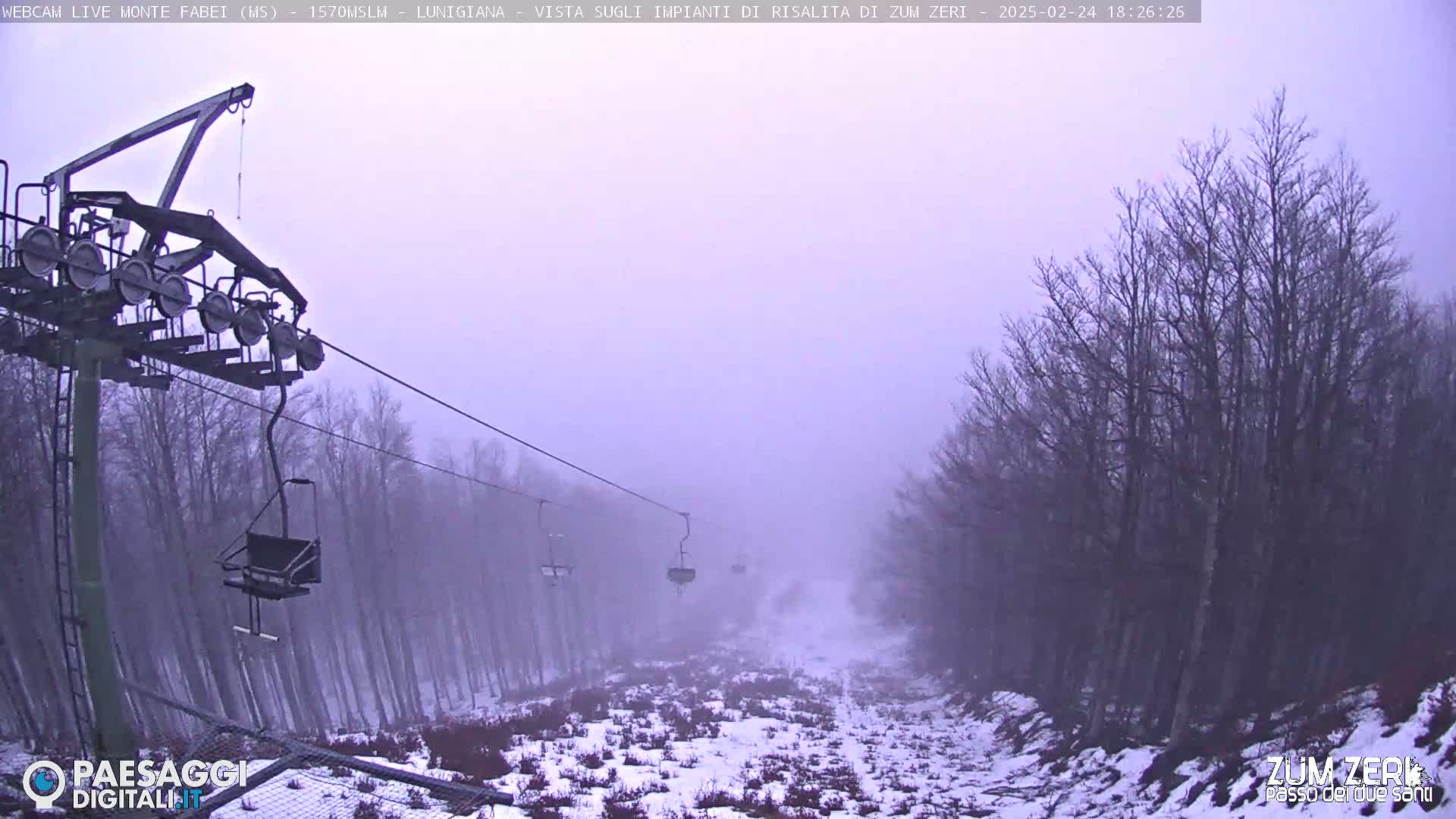 A chairlift ascends a snowy, foggy mountain slope lined with leafless trees.
