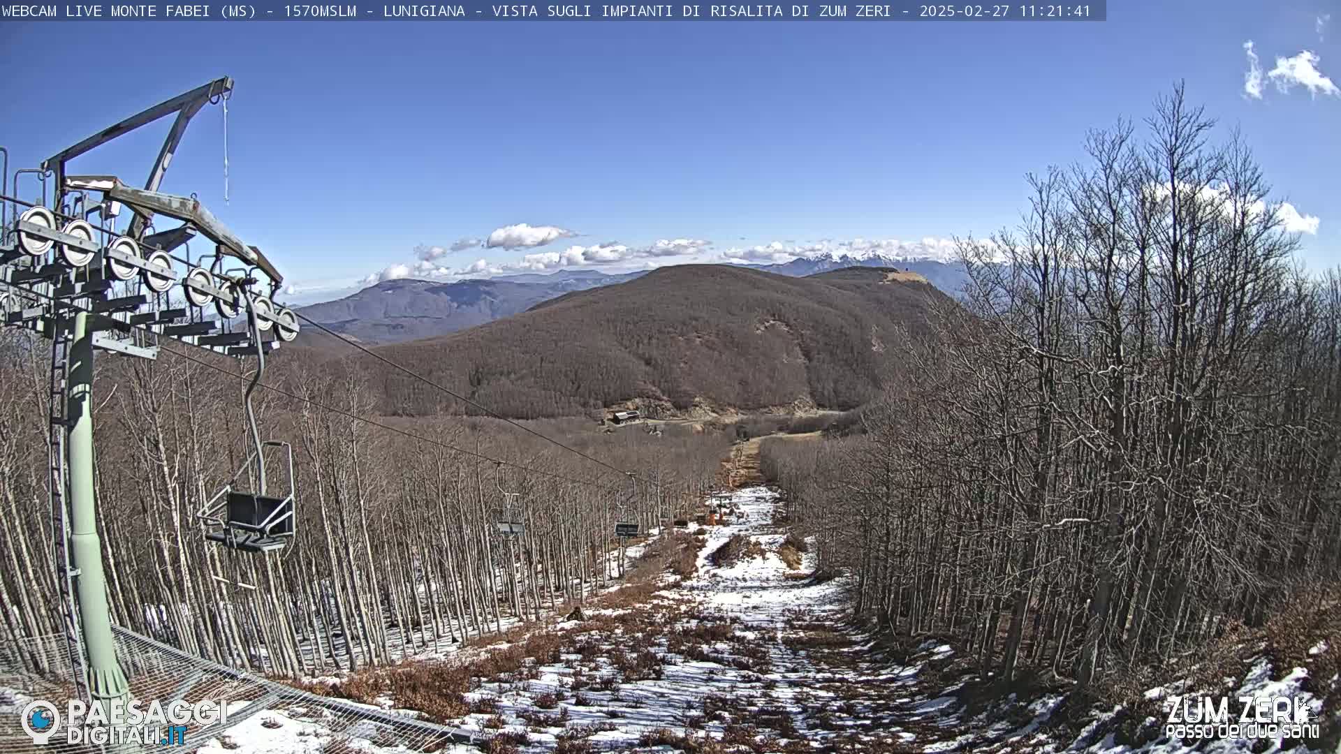 A chairlift ascends a mountainside sparsely covered in snow under a clear blue sky with a few clouds.