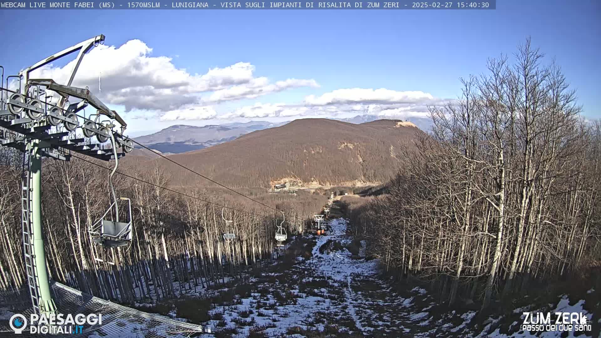 A chairlift ascends a mountainside covered in leafless trees and patches of snow under a partly cloudy sky.
