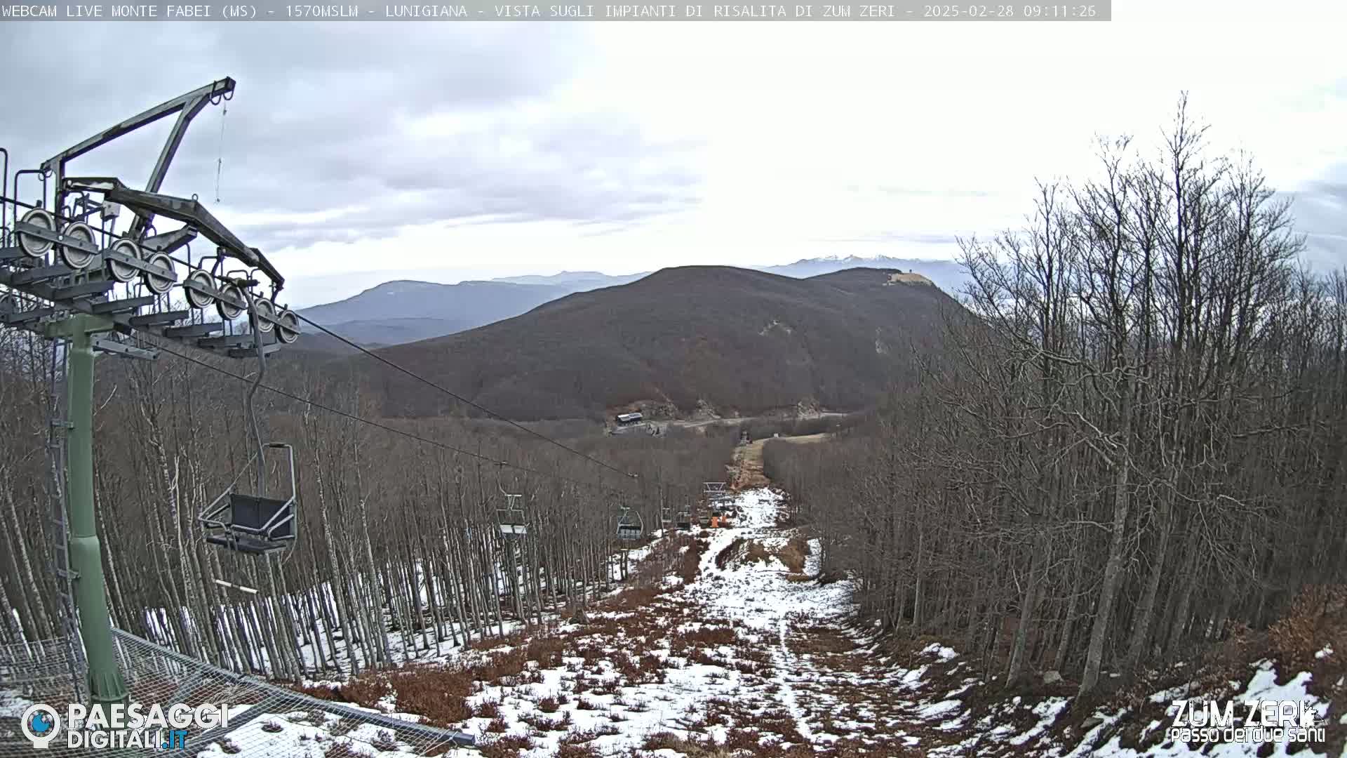 A chairlift ascends a snow-dusted mountainside under a cloudy sky, passing through a sparse, leafless forest.
