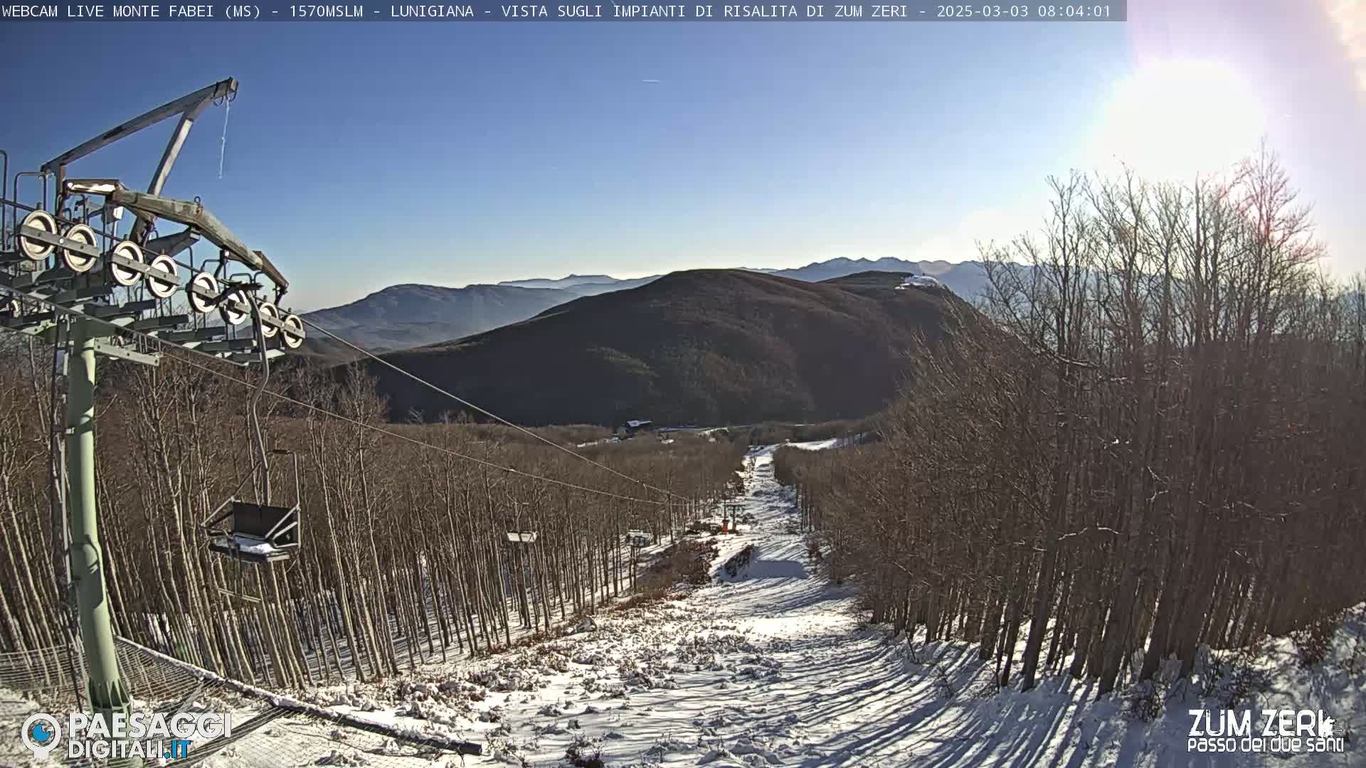 A chairlift ascends a snowy mountain slope lined with bare trees under a clear, sunny sky.