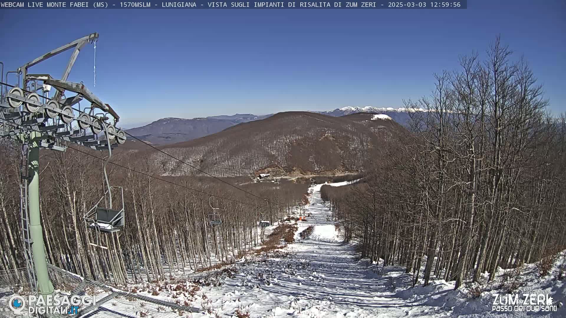 A chairlift ascends a snow-covered ski slope through a leafless forest on a sunny day.