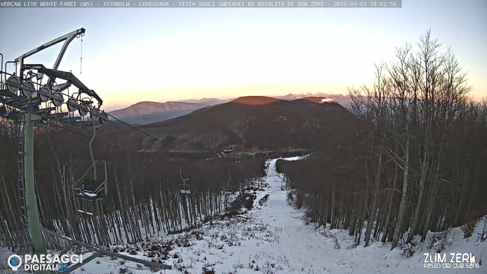 A ski lift descends a snow-covered mountain slope at sunset, surrounded by bare trees.