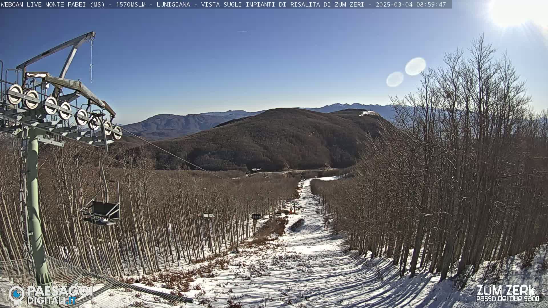 A ski lift ascends a snowy mountainside on a sunny day, surrounded by leafless trees.