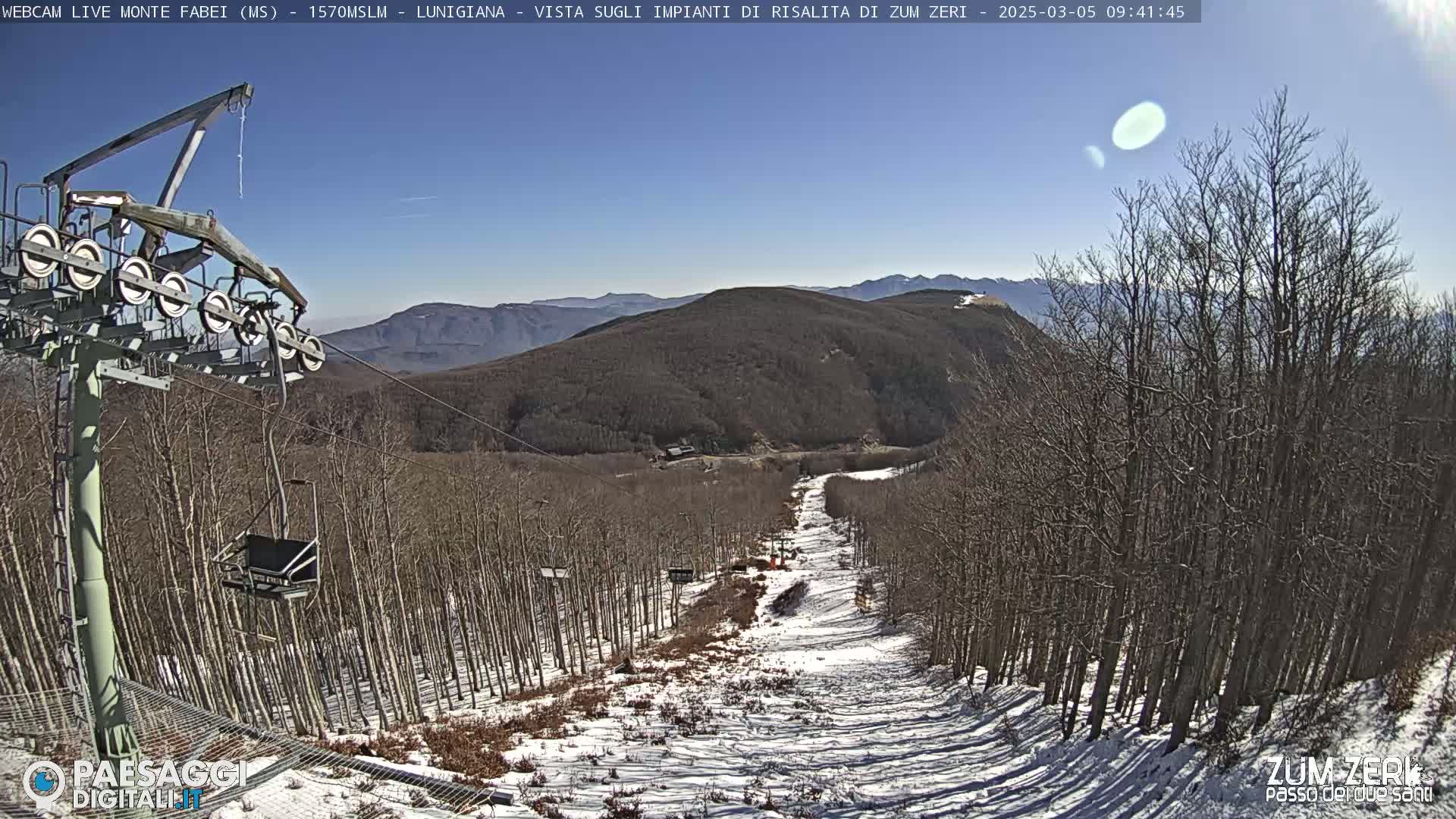 A chairlift ascends a snowy ski slope through a forest of leafless trees on a sunny day.
