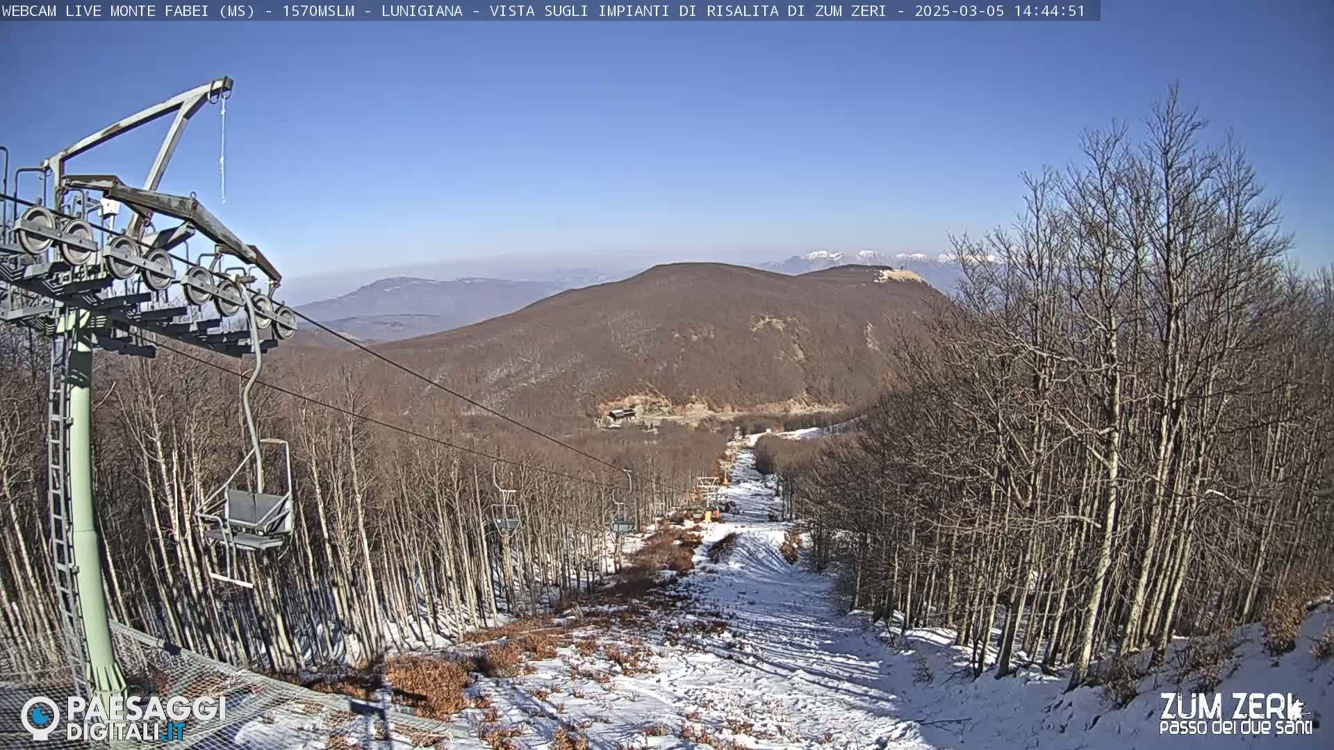A chairlift ascends a snow-dusted mountain slope lined with bare trees under a clear blue sky.