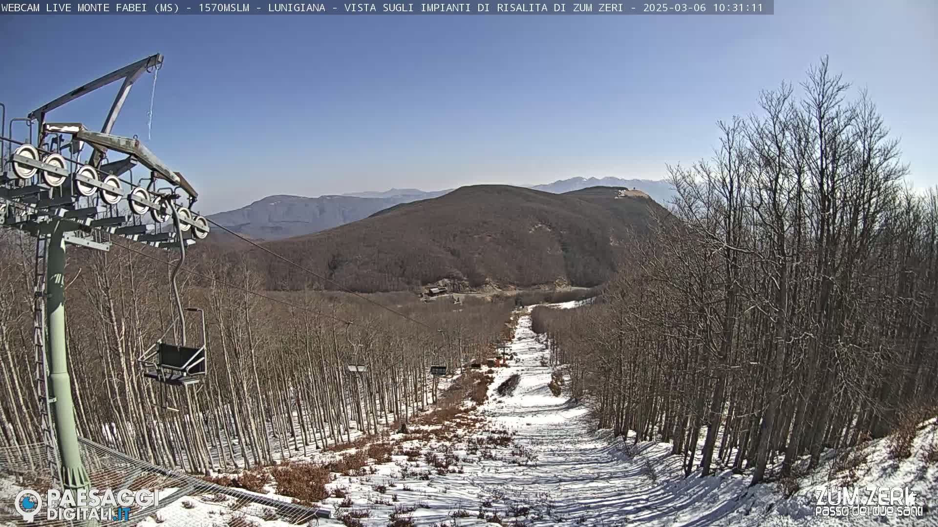 A chairlift ascends a snowy mountain slope lined with bare trees under a clear blue sky.