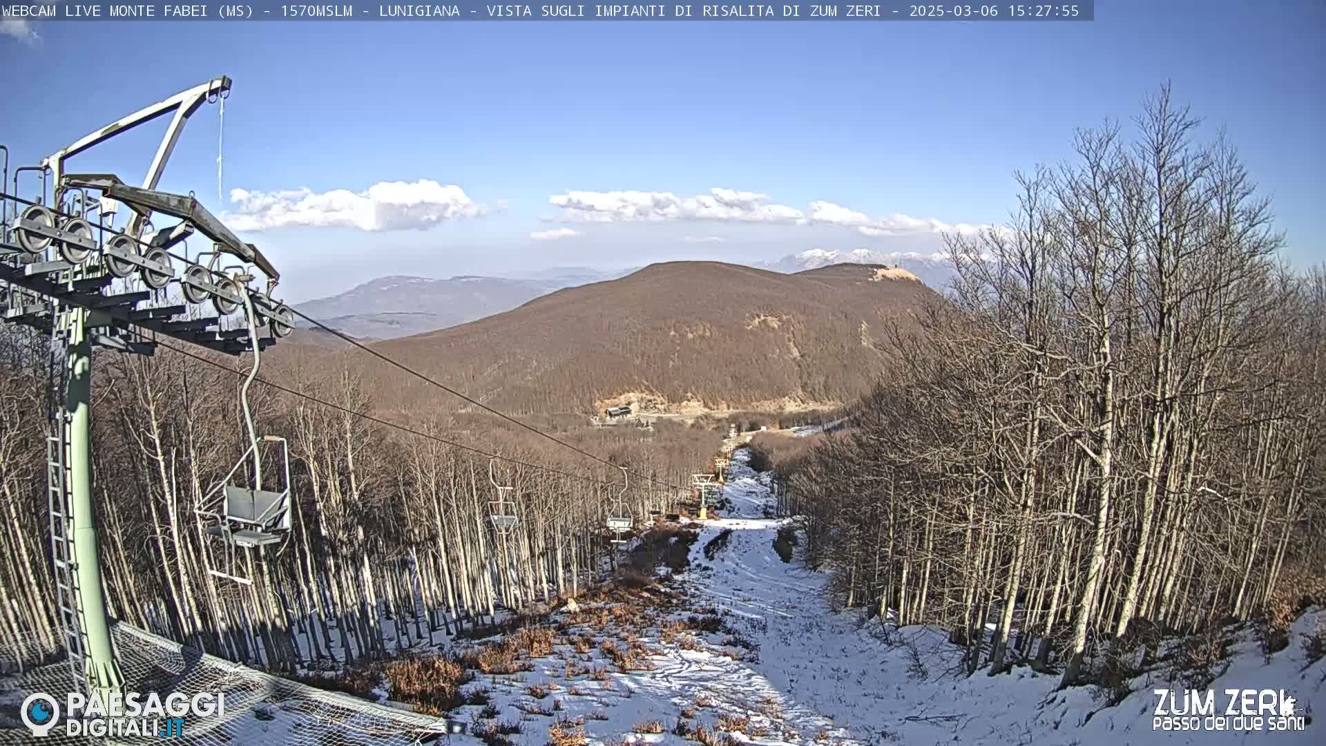 A chairlift ascends a snow-dusted mountainside, under a mostly sunny sky with a few scattered clouds, passing through a forest of bare, winter trees.