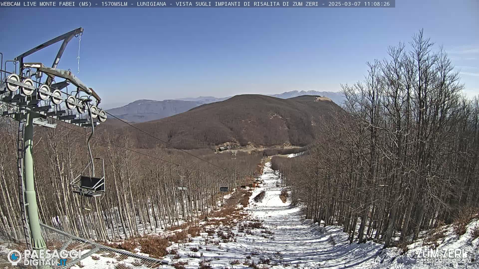 A ski lift chair ascends a snowy mountain slope surrounded by leafless trees under a clear blue sky.