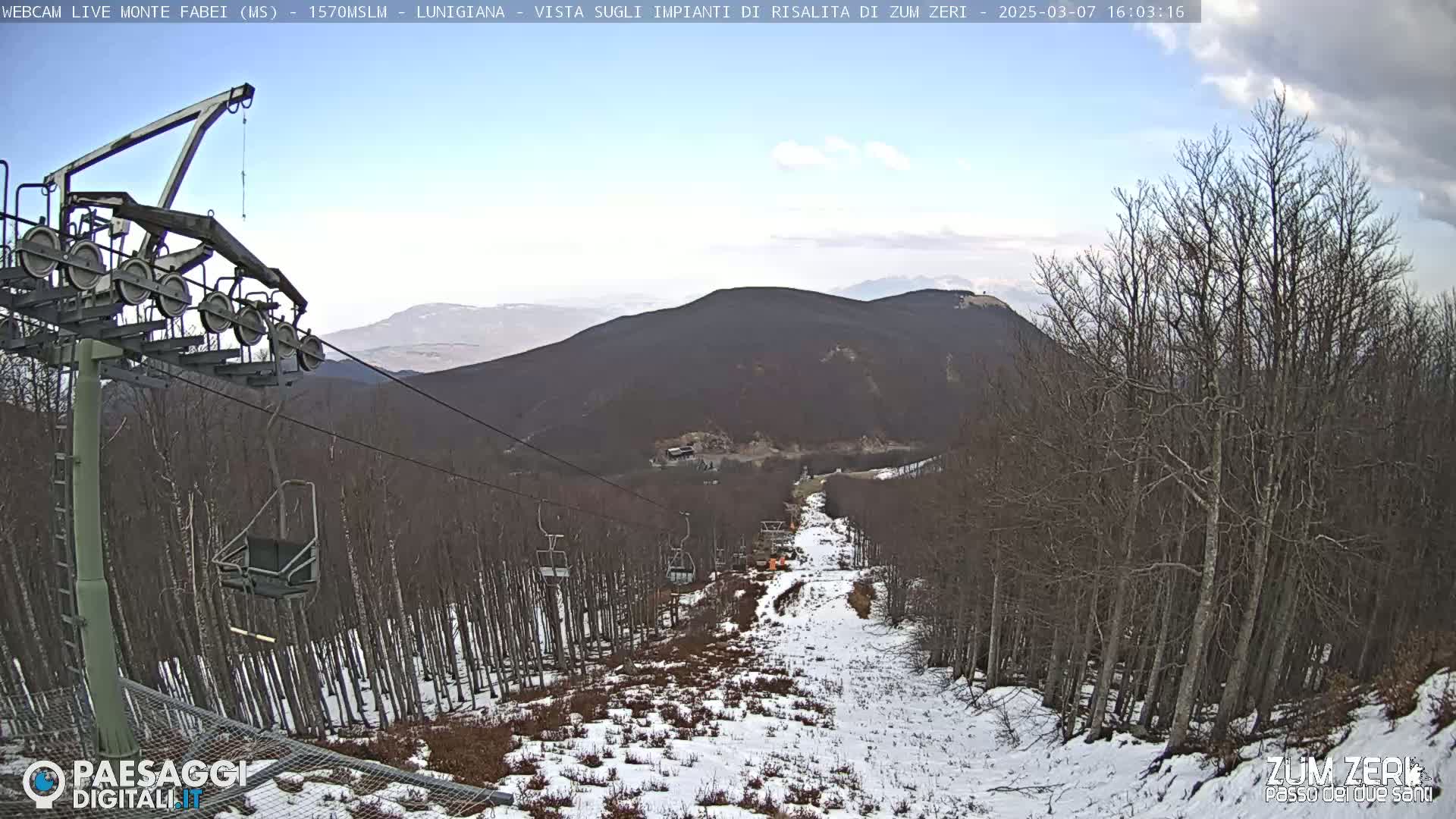 A chairlift ascends a snow-dusted mountain slope lined with leafless trees under a partly cloudy sky.