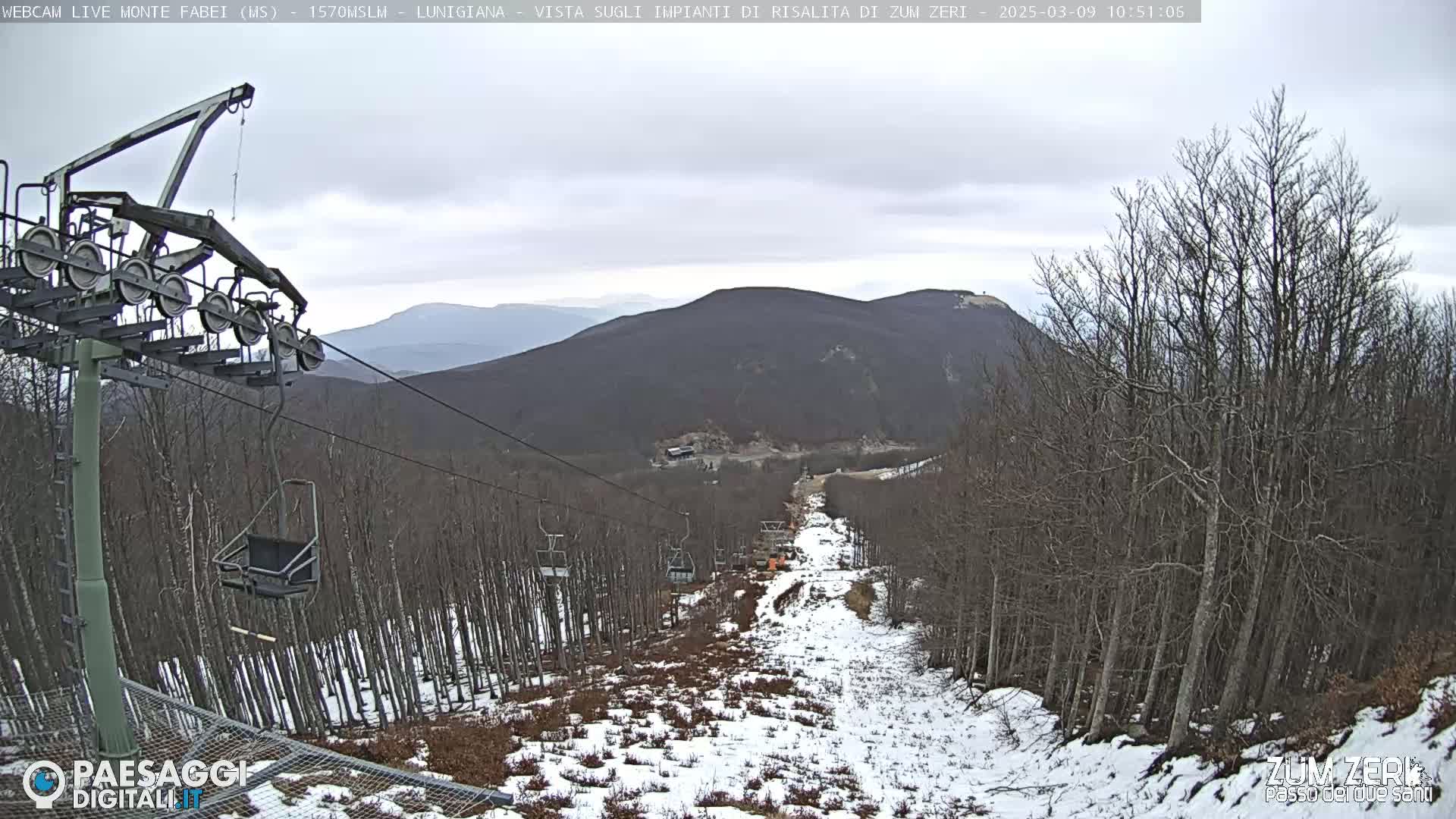 A chairlift ascends a snowy mountain slope amongst bare, leafless trees under an overcast sky.