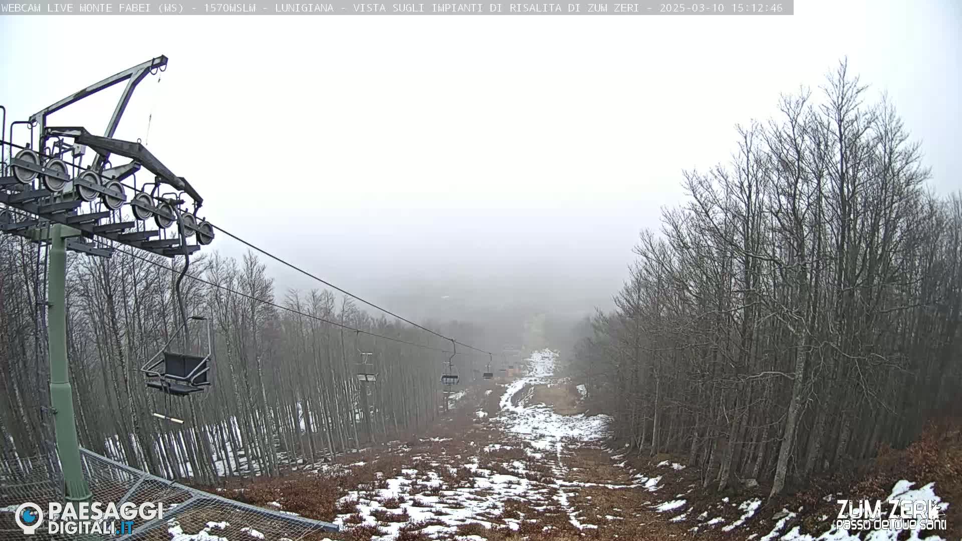 A foggy view down a ski slope with a chairlift and bare trees partially covered in snow.