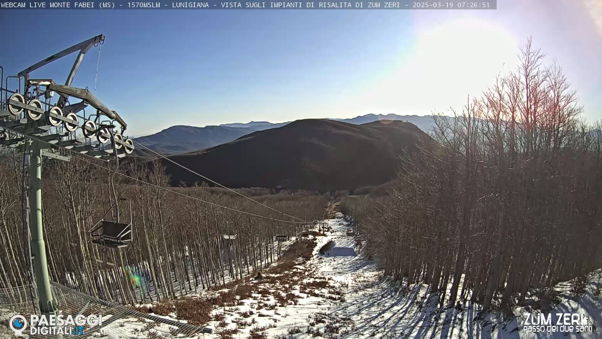 A chairlift ascends a snowy mountainside lined with bare trees under a sunny sky.