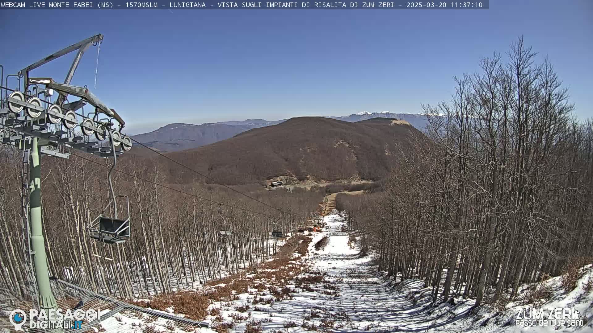 A ski lift chair ascends a snow-dusted mountain slope, under a clear blue sky, surrounded by leafless trees.