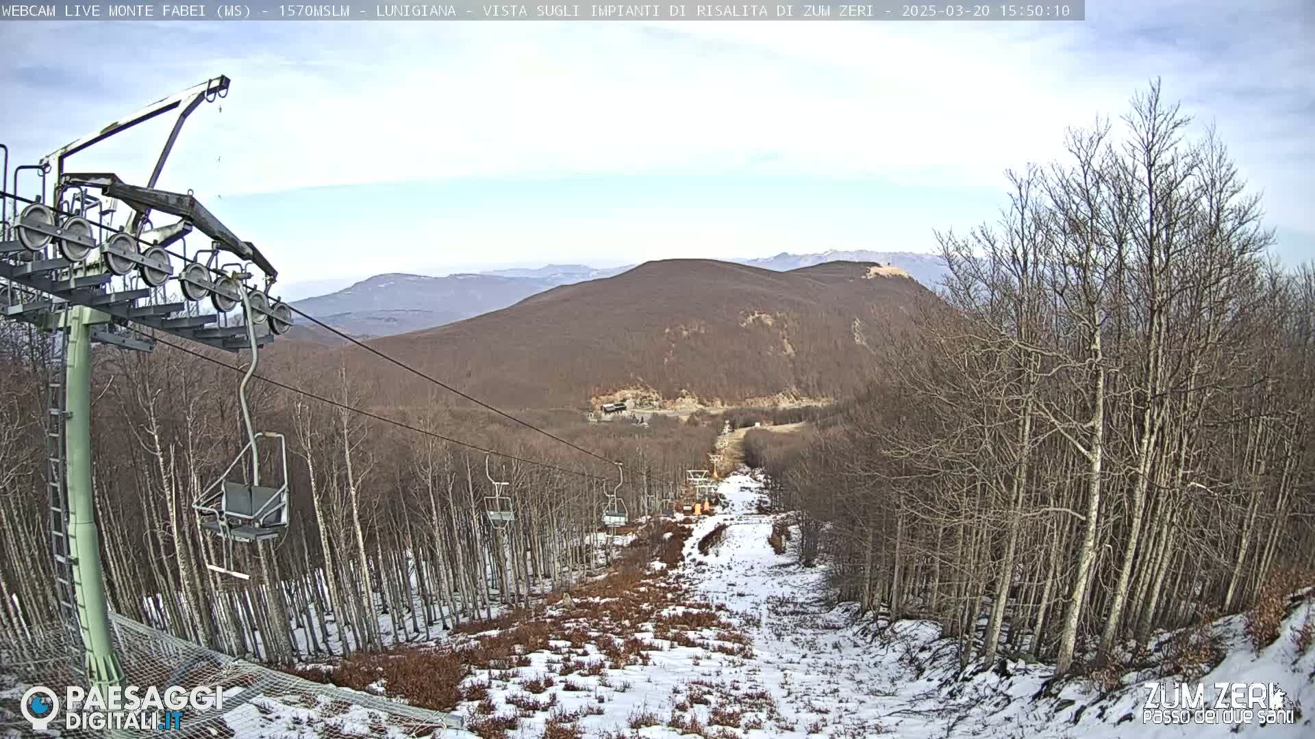 A chairlift ascends a snowy mountain slope lined with bare trees under a mostly cloudy sky.