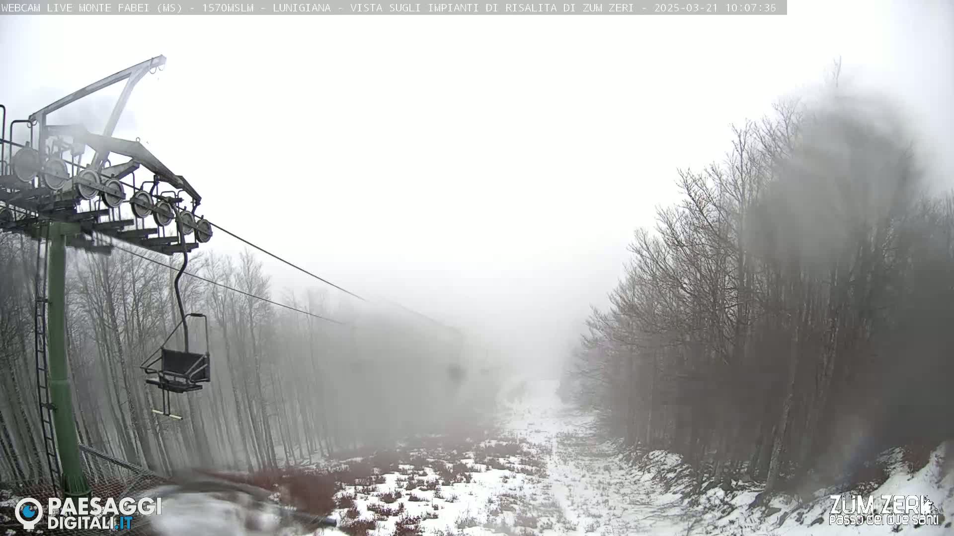 A ski lift chair ascends a snowy, foggy mountain slope lined with bare trees.
