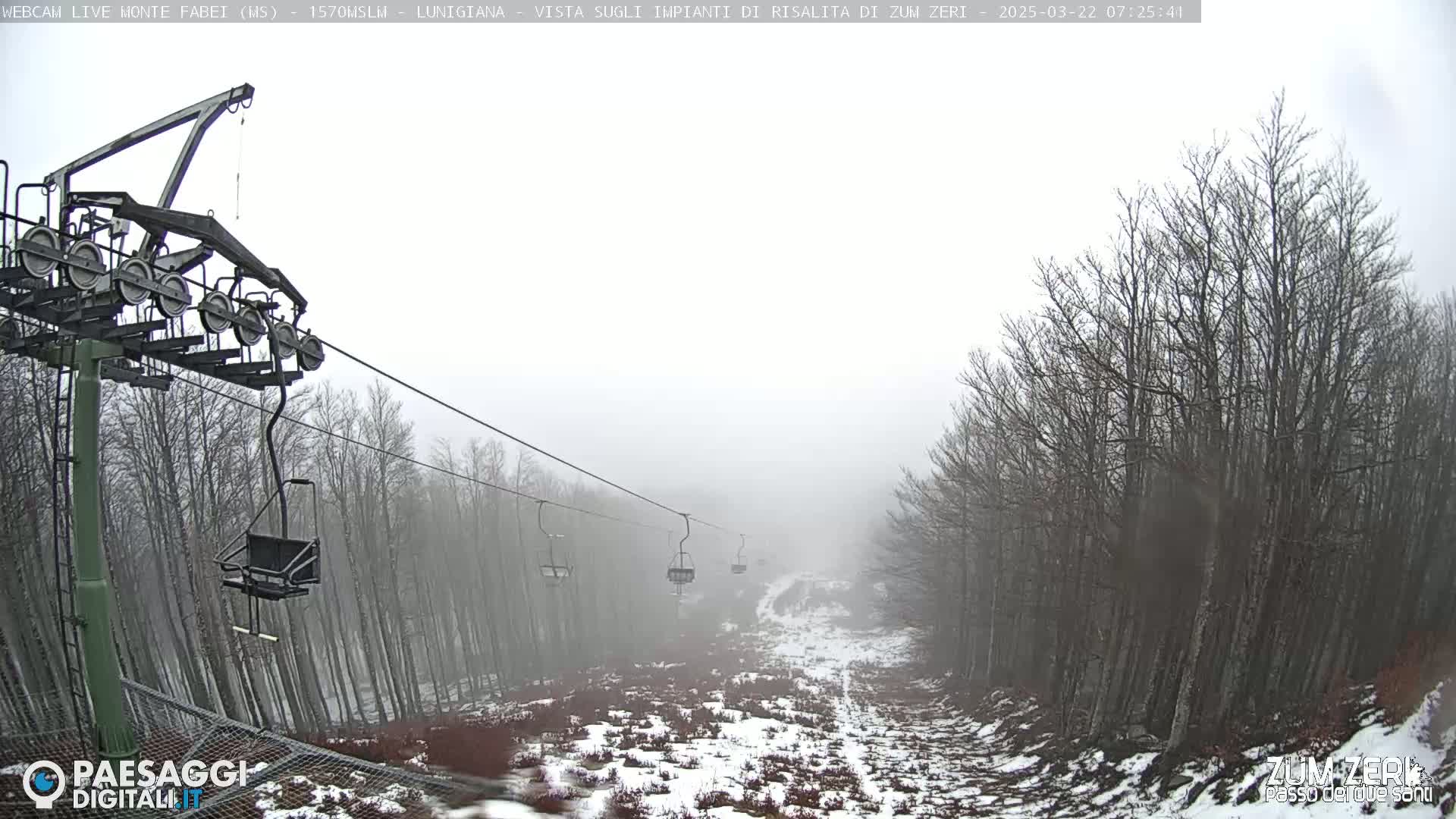 A chairlift ascends a foggy mountain slope lined with leafless trees and patches of snow.