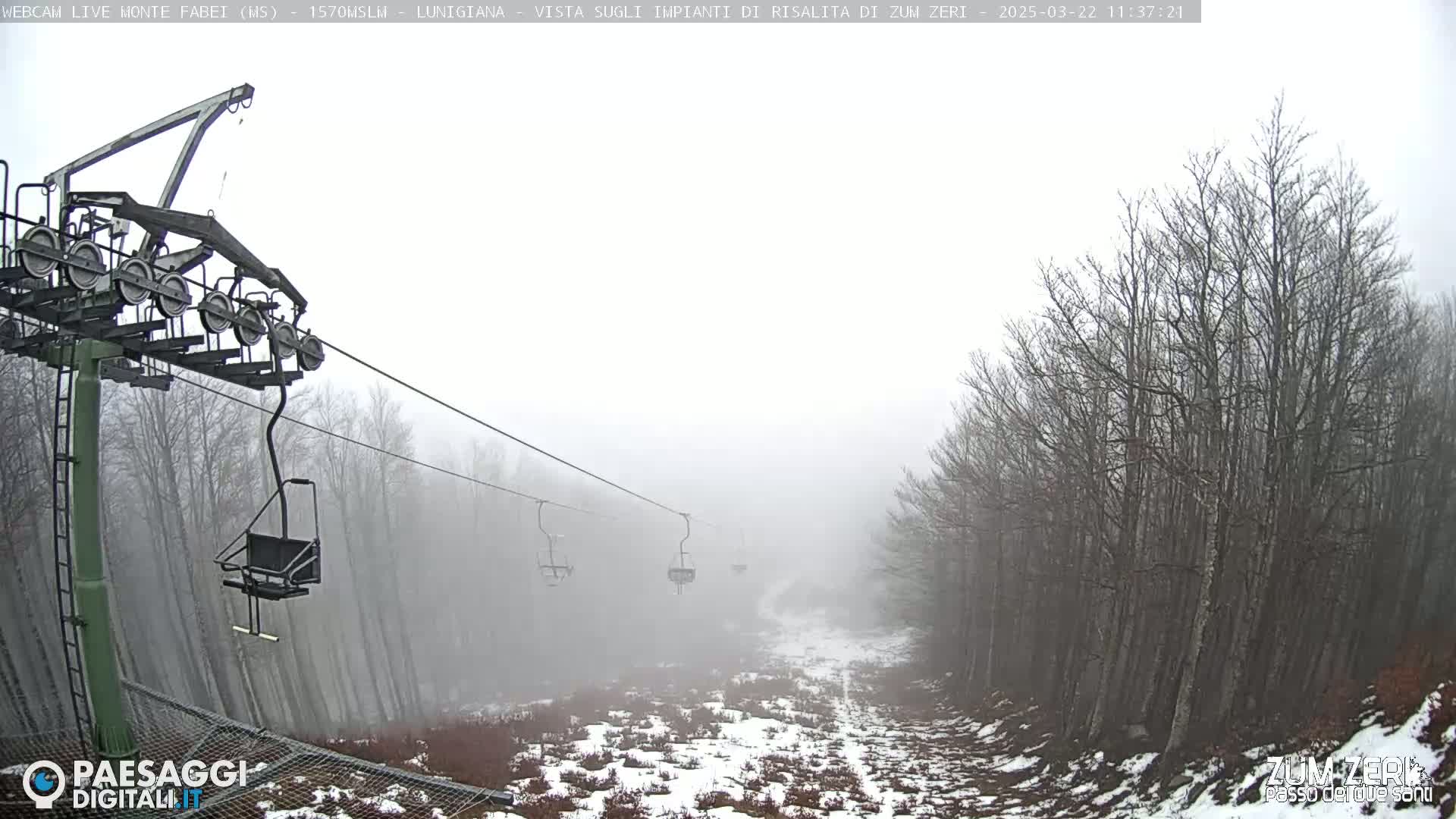 A chairlift ascends a foggy mountain slope lined with leafless trees and patches of snow.