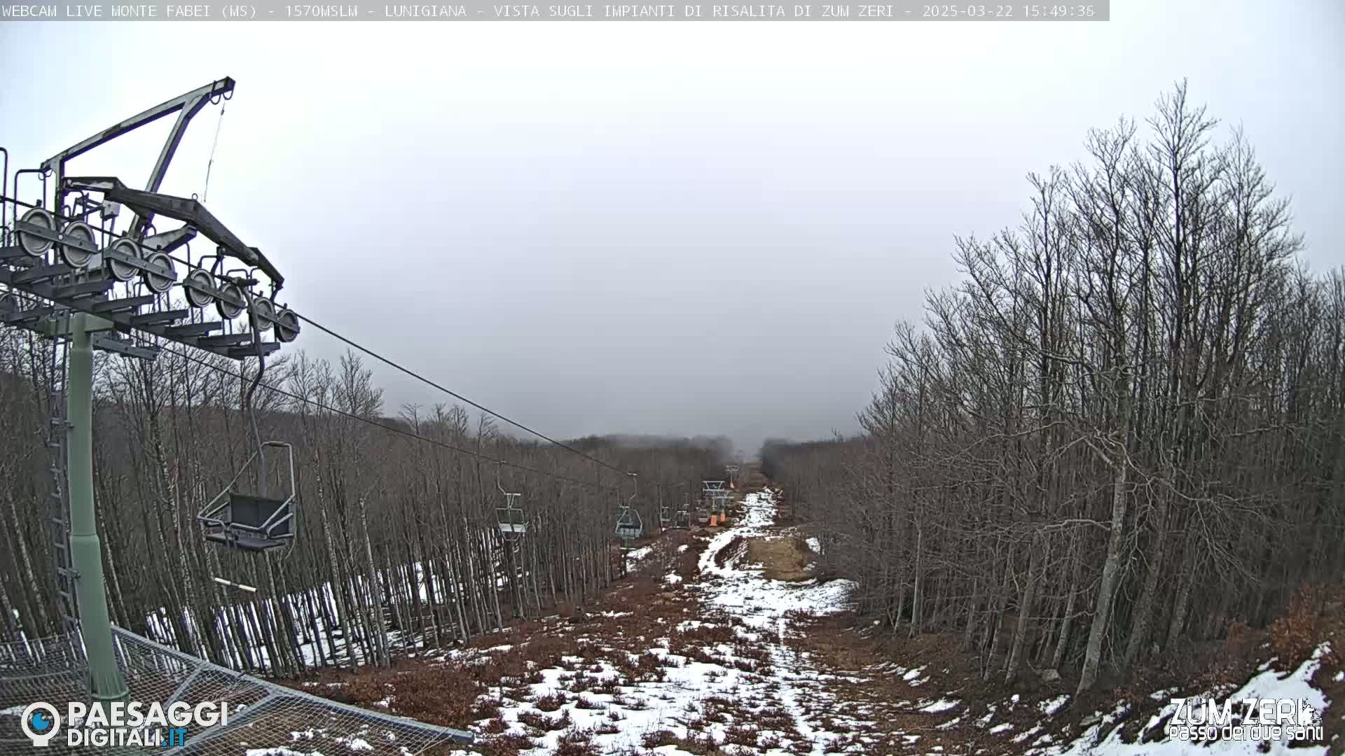 A chairlift extends down a snowy, barren hillside lined with leafless trees under an overcast sky.