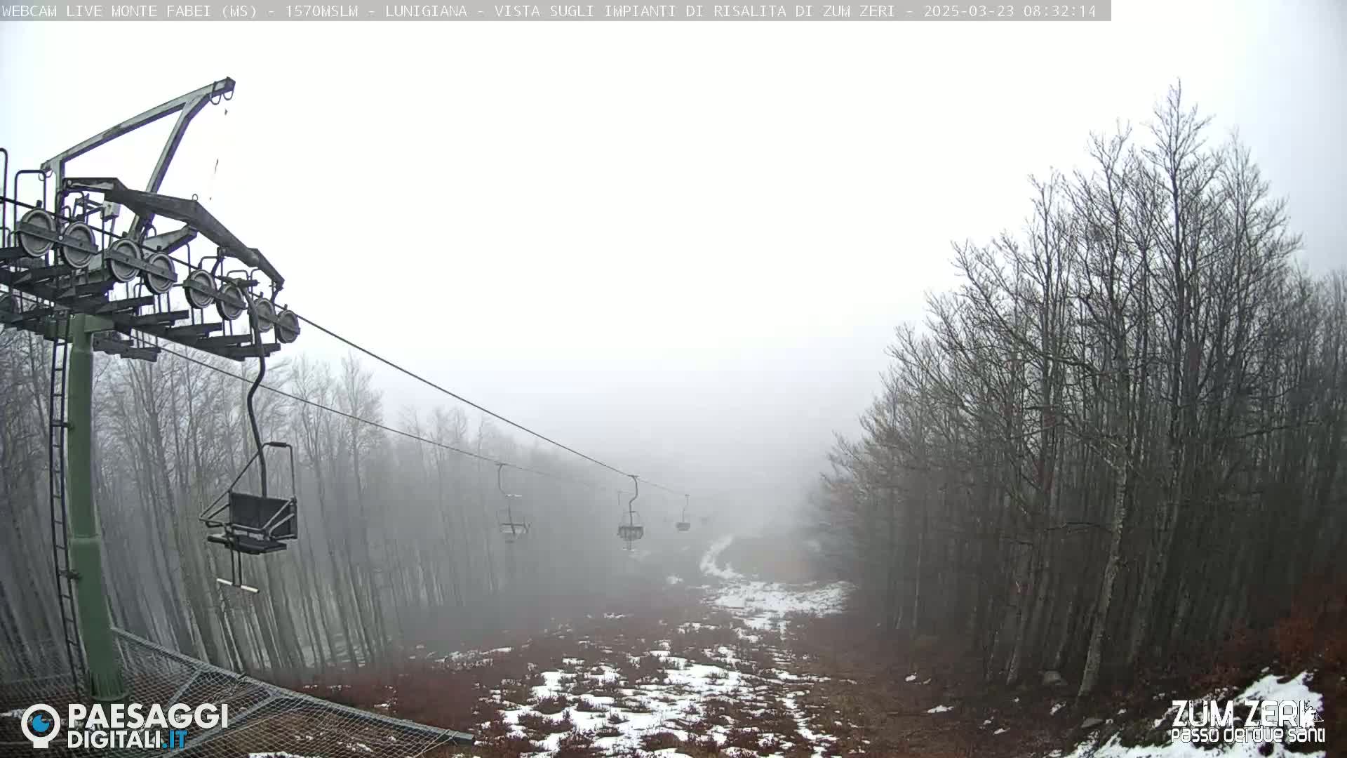 A foggy view of a chairlift ascending through a sparsely snow-covered forest of bare trees.
