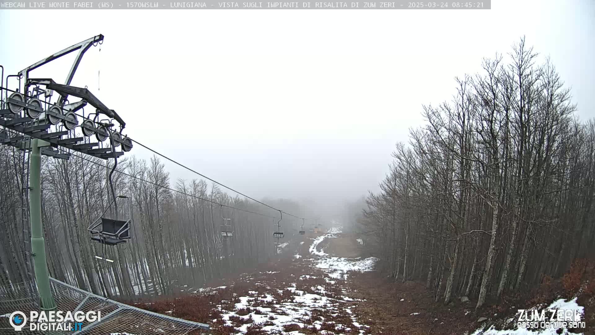 A foggy view of a chairlift ascending a sparsely snow-covered hill lined with leafless trees.