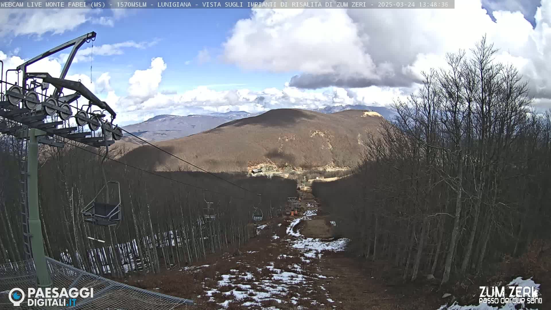 A ski lift ascends a mountainside under a partly cloudy sky, with patches of snow visible on the ground and bare trees lining the slopes.