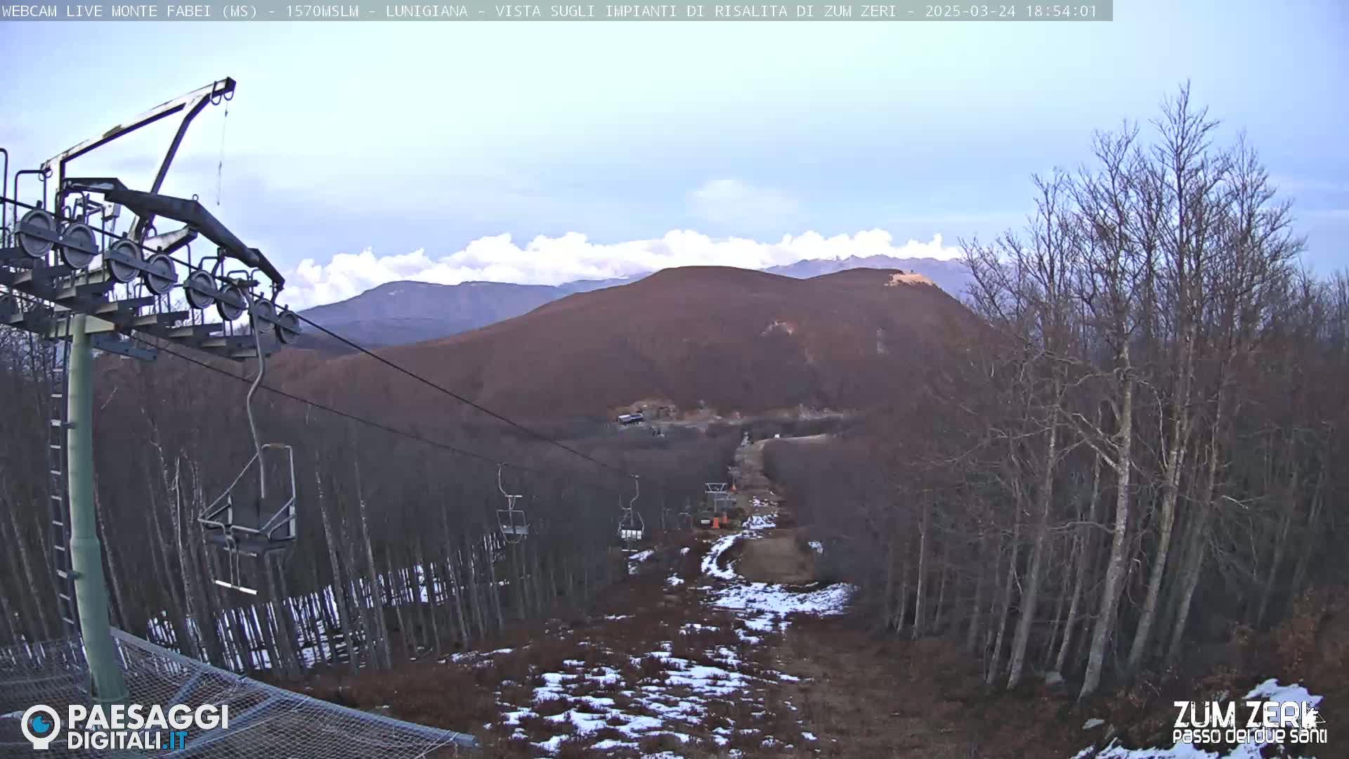 A chairlift ascends a mountainside through a mostly leafless forest with patchy snow on the ground under a partly cloudy sky.