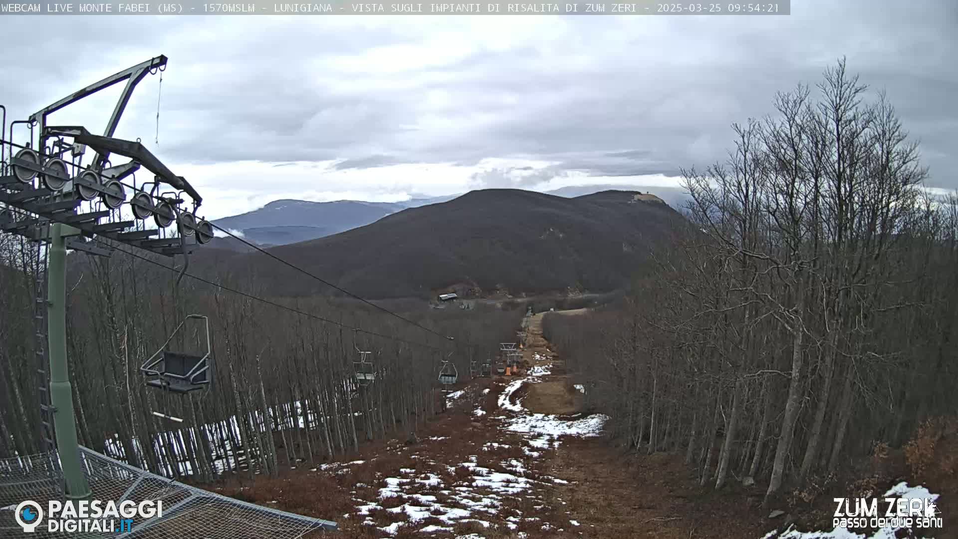 A chairlift ascends a sparsely snow-covered mountainside under a cloudy sky.