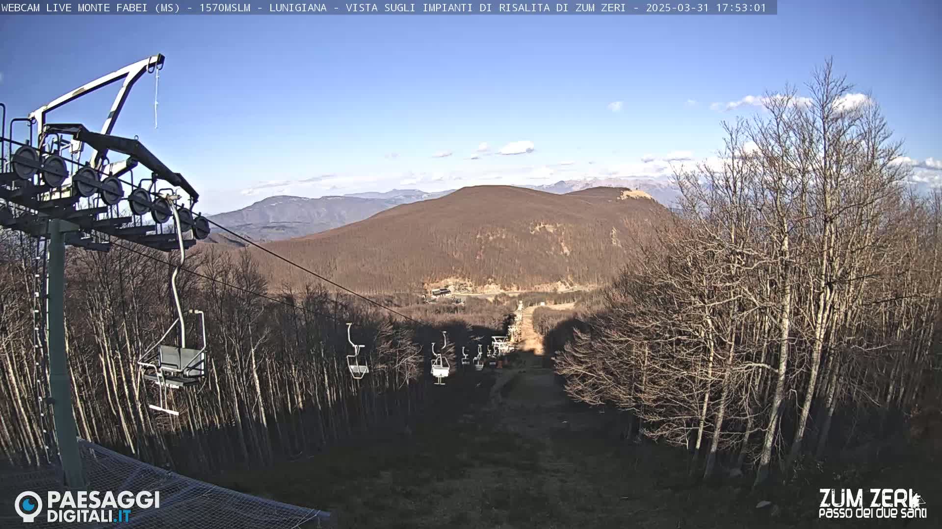 A chairlift ascends a mountainside covered in leafless trees under a mostly clear, sunny sky.