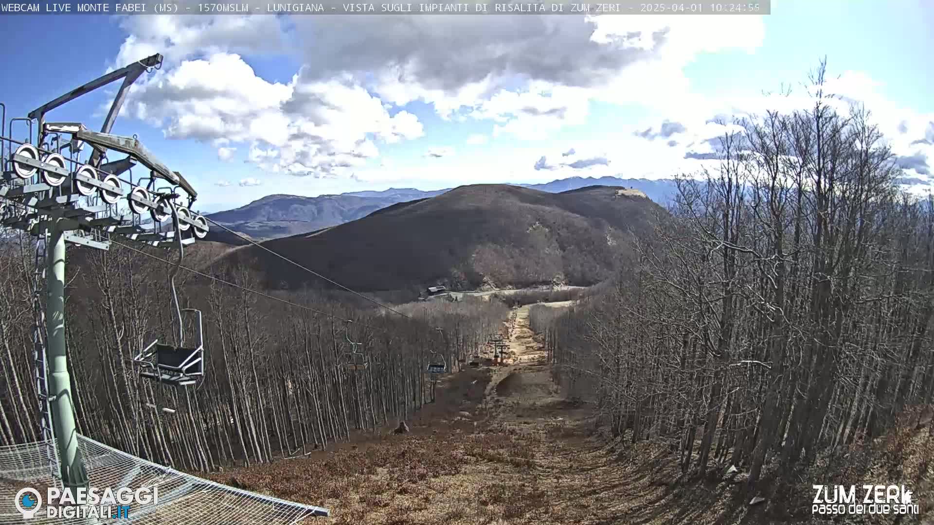 A chairlift ascends a mountainside covered in bare, brown trees under a partly cloudy sky.
