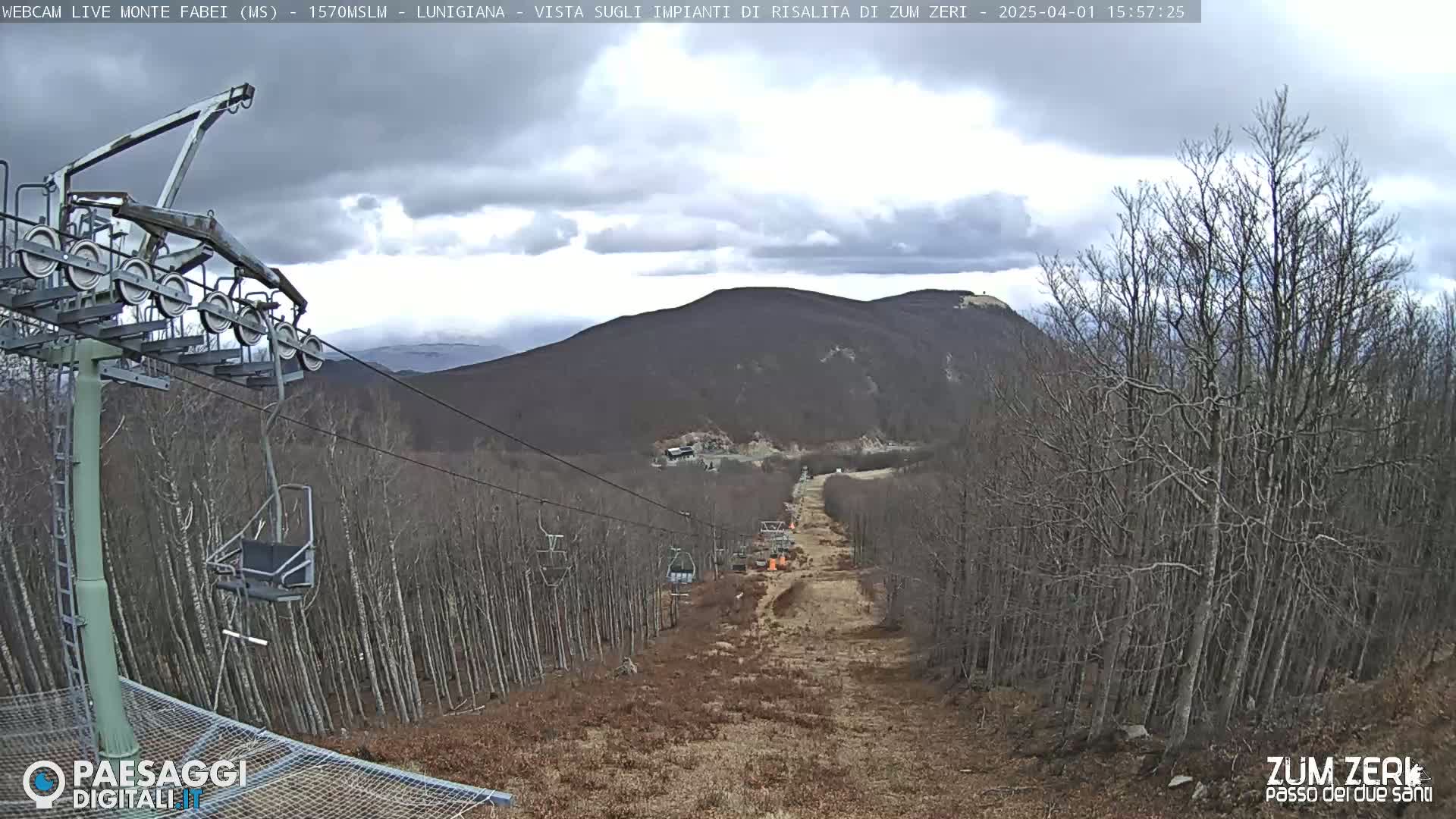 A chairlift ascends a mountainside under an overcast sky, passing through a barren, leafless forest.