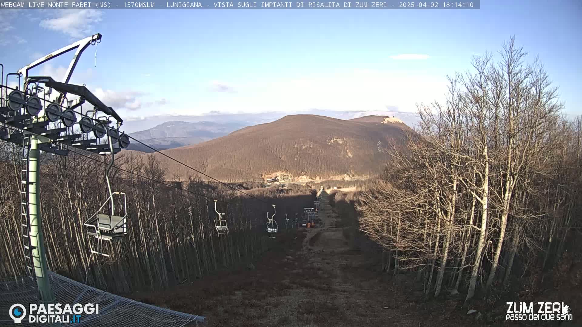 A chairlift ascends a mountainside under a partly cloudy sky, passing through bare, winter trees toward a distant mountain peak.