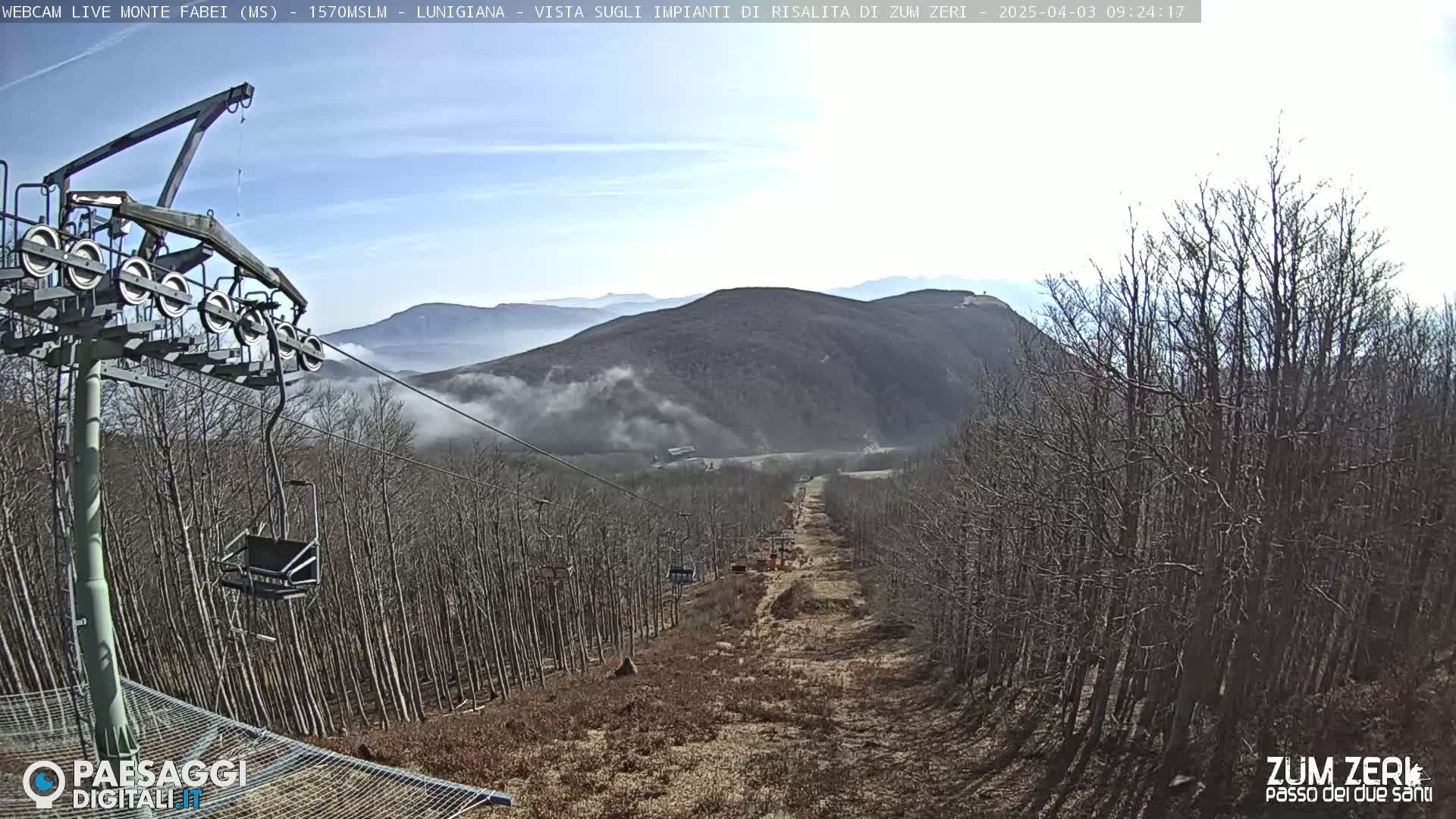 A chairlift ascends a mountainside through leafless trees under a mostly clear, bright sky with some low-lying fog.