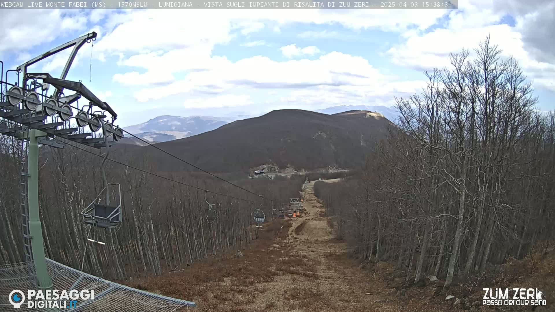 A chairlift ascends a mountainside under a partly cloudy sky, passing through a sparsely wooded area with brown undergrowth.