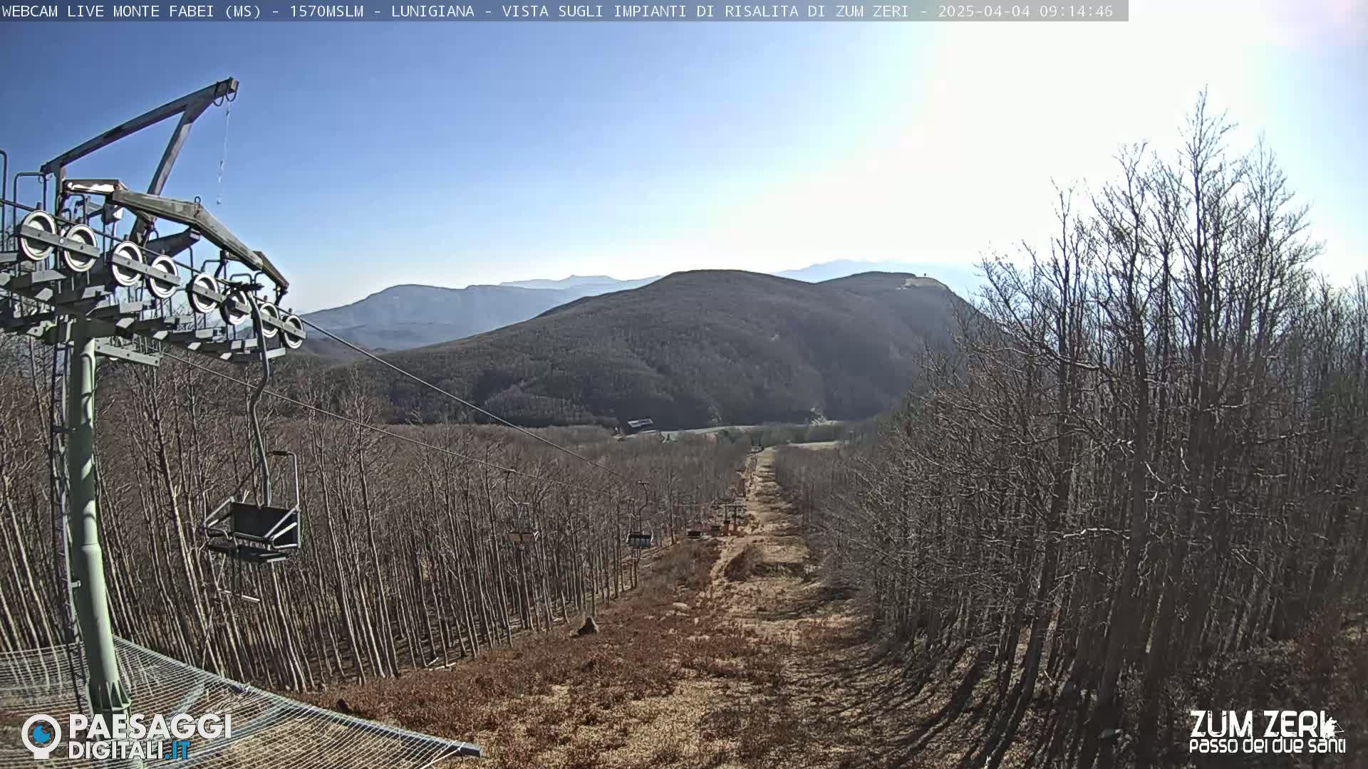 A ski lift ascends a hill covered in leafless trees under a clear, sunny sky.