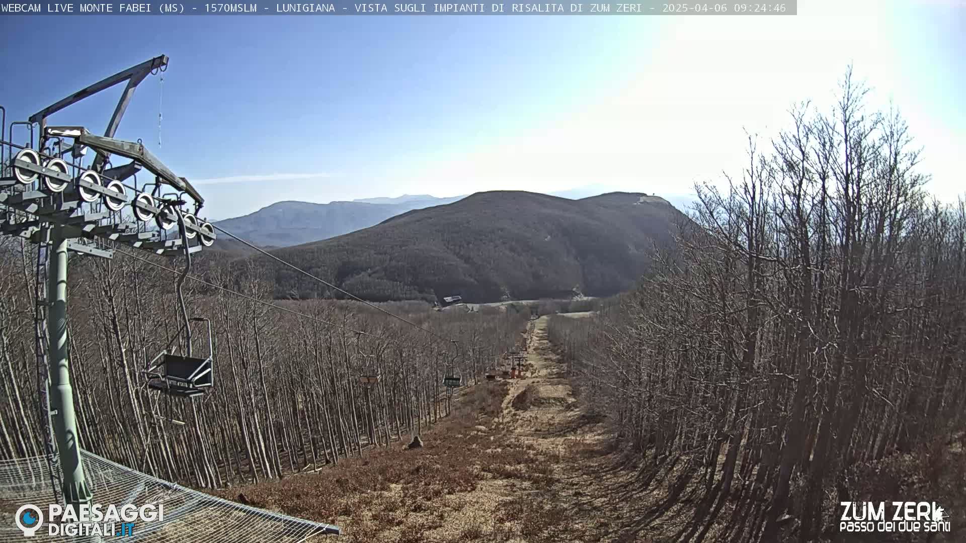 A chairlift ascends a mountainside through a forest of bare trees under a clear blue sky.