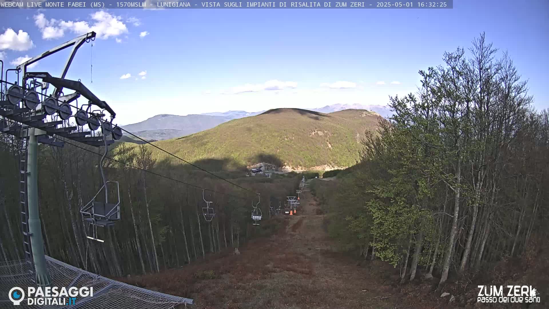 A mostly clear sky overlooks a chairlift ascending a mountainside covered in budding trees and brown undergrowth.