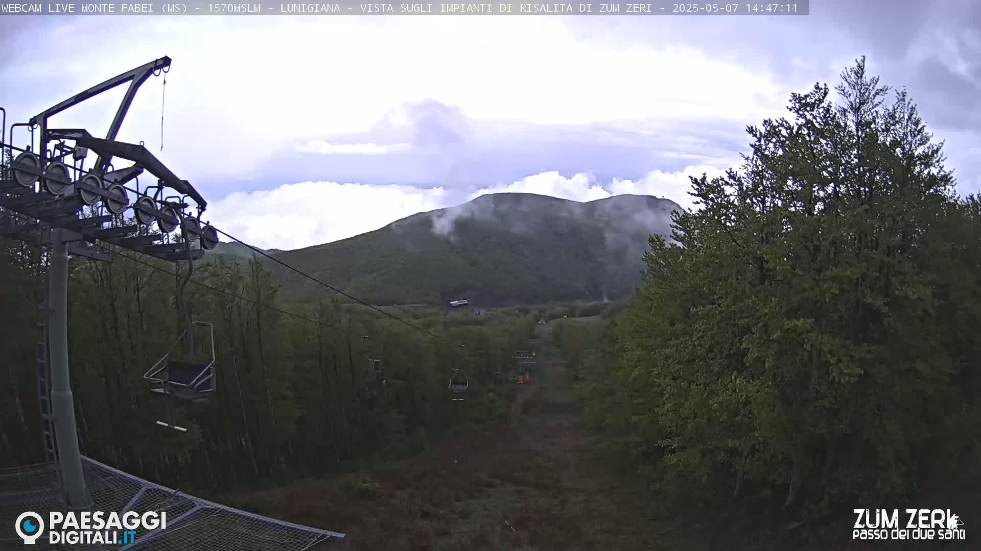 A chairlift ascends a mountain partially obscured by clouds, passing through a lush green forest.
