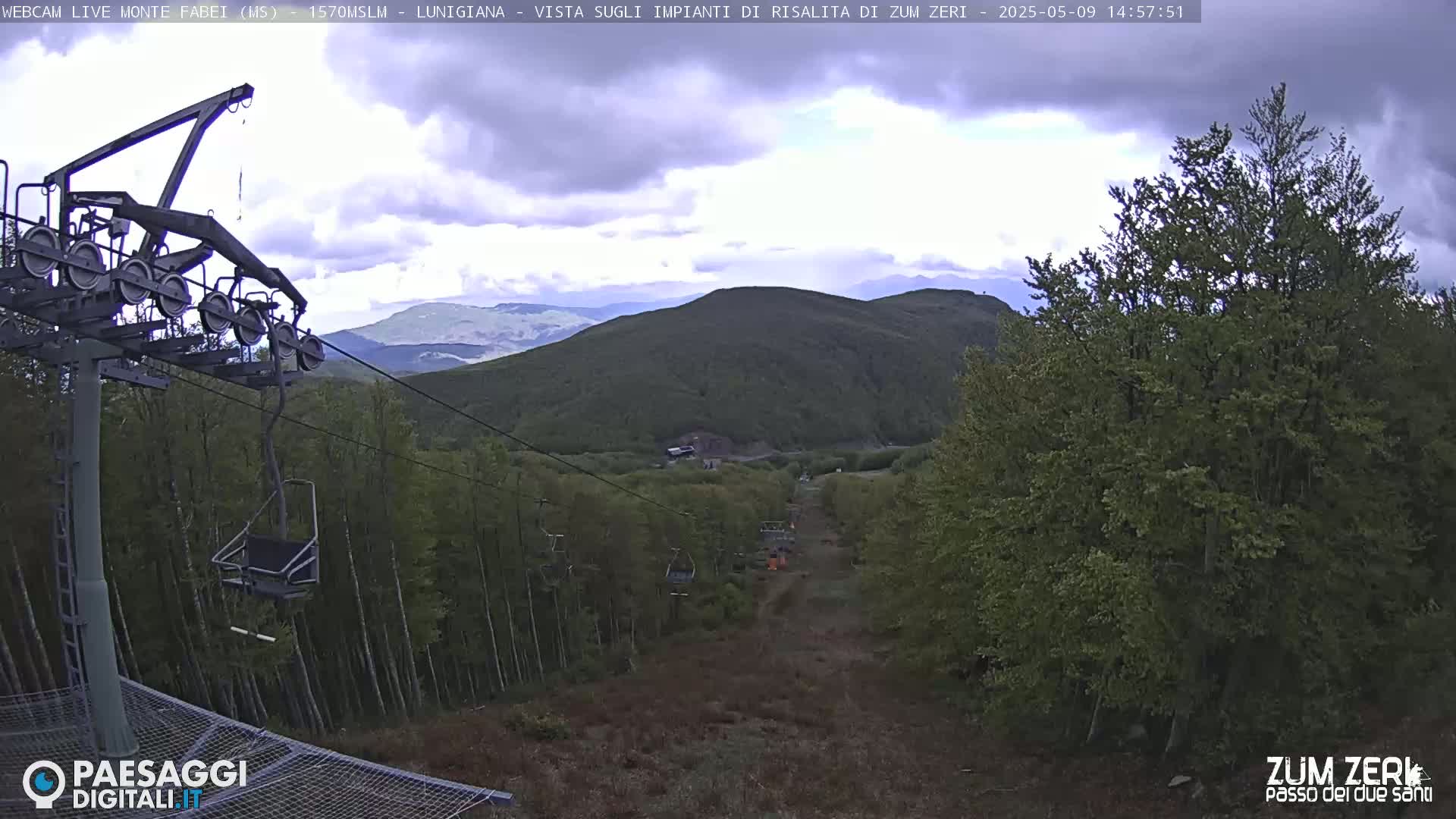 A chairlift ascends a mountainside through a forest under a partly cloudy sky.