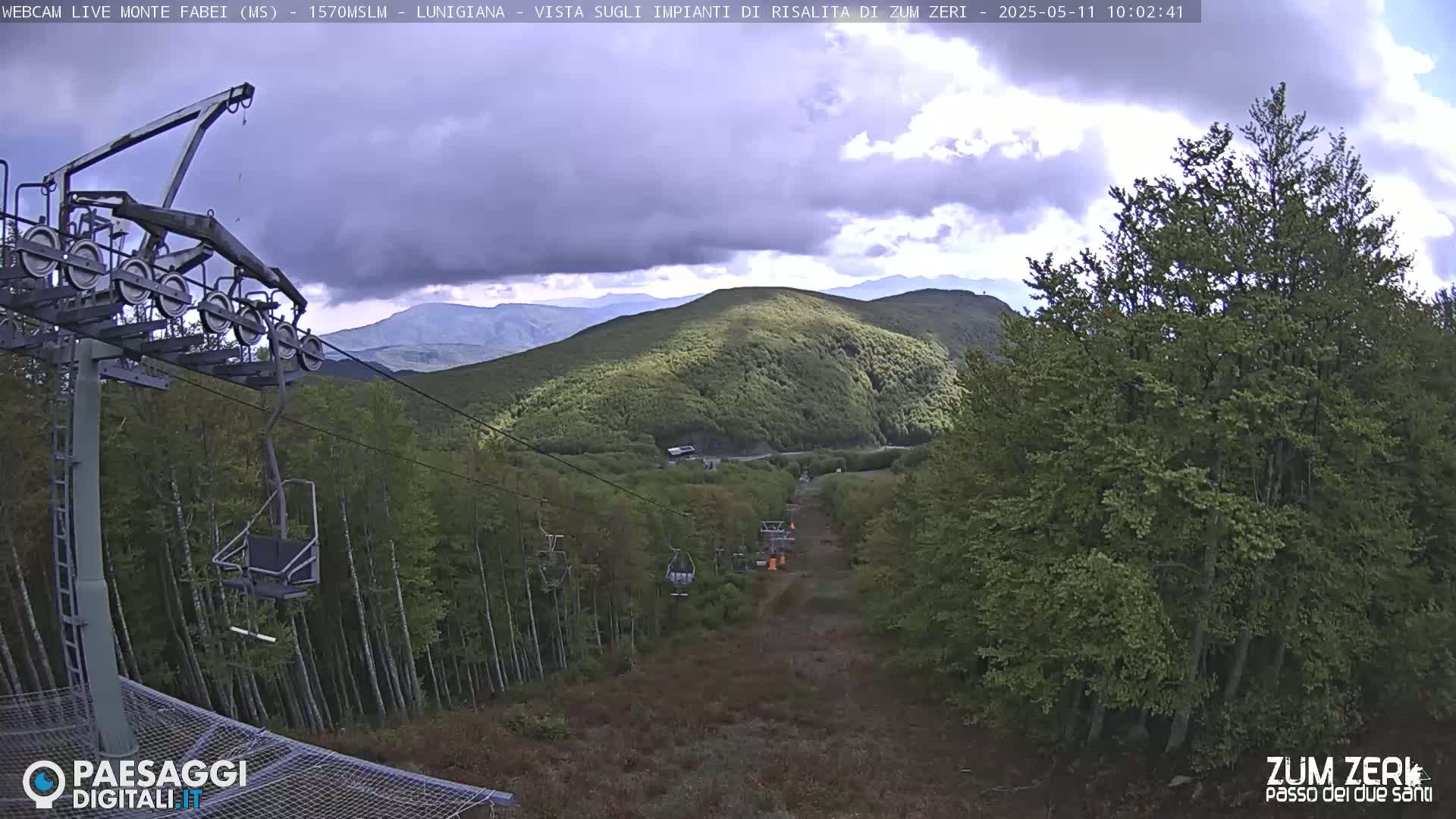 A chairlift ascends a mountainside covered in lush green trees under a partly cloudy sky.