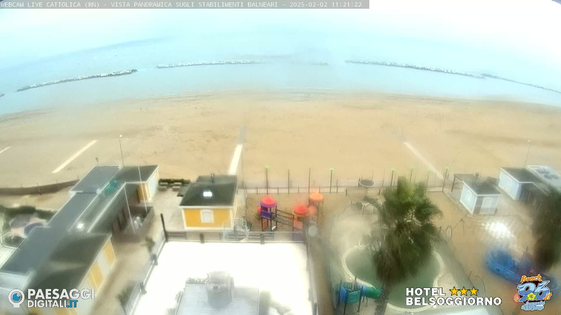 A hazy, overcast day at a beach, showing a sandy shore, calm water, breakwaters, beachside buildings, a playground, and a swimming pool.
