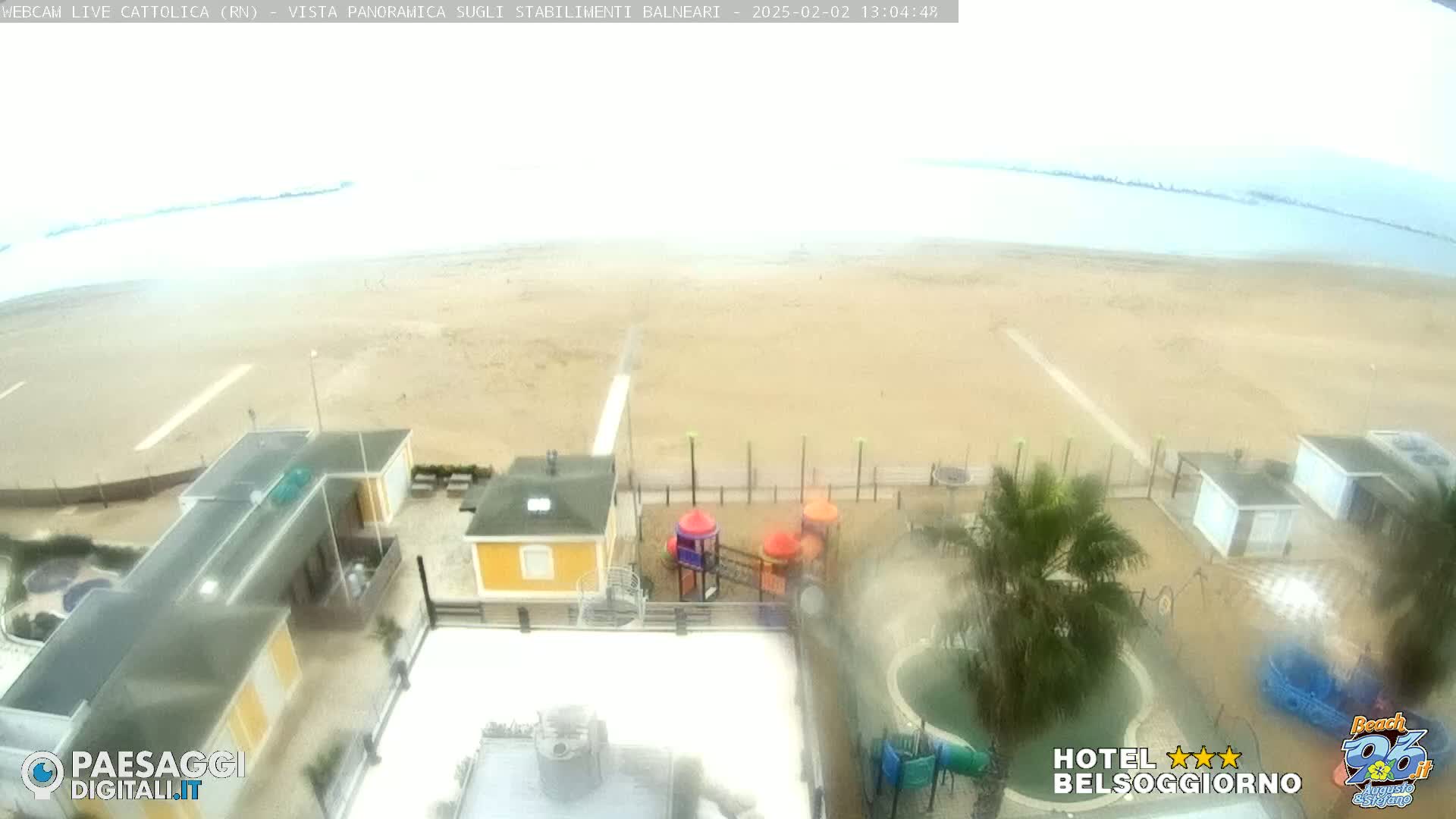 A hazy, overcast view of a beach with several small buildings, a playground, and a pool, seen from an elevated vantage point.